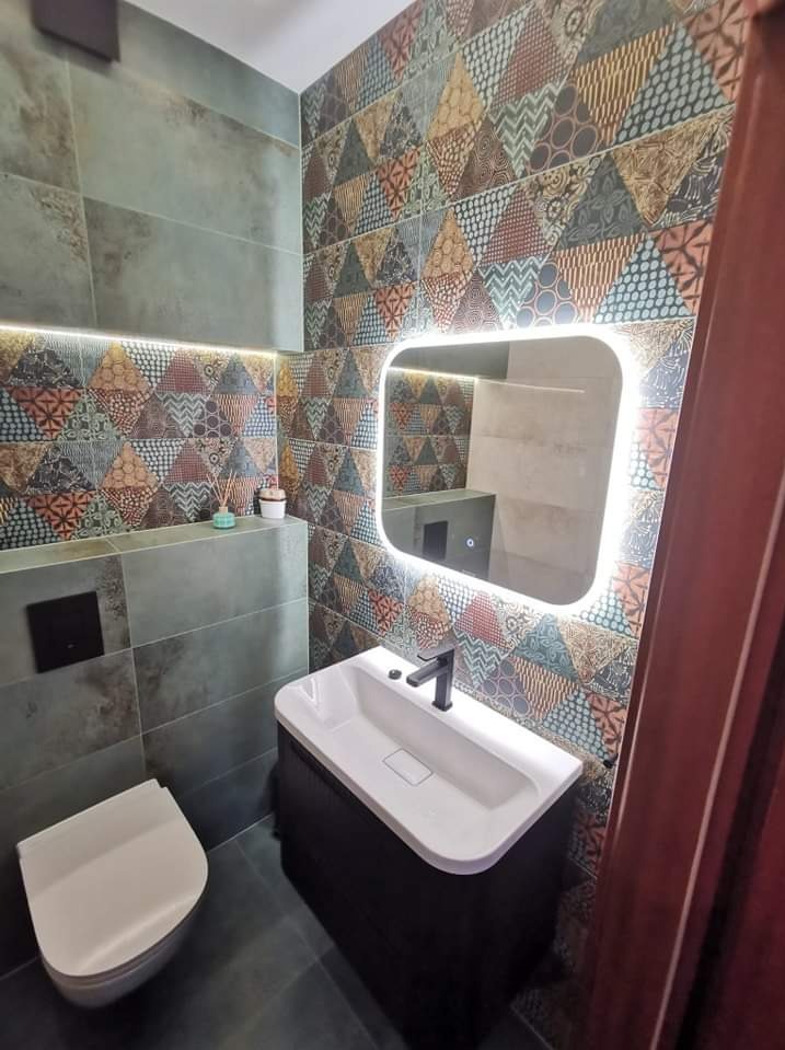 Nowoczesna łazienka z patchworkowymi płytkami na ścianie, podświetlane lustro nad umywalką z czarną armaturą, szara toaleta i półka z dekoracjami.