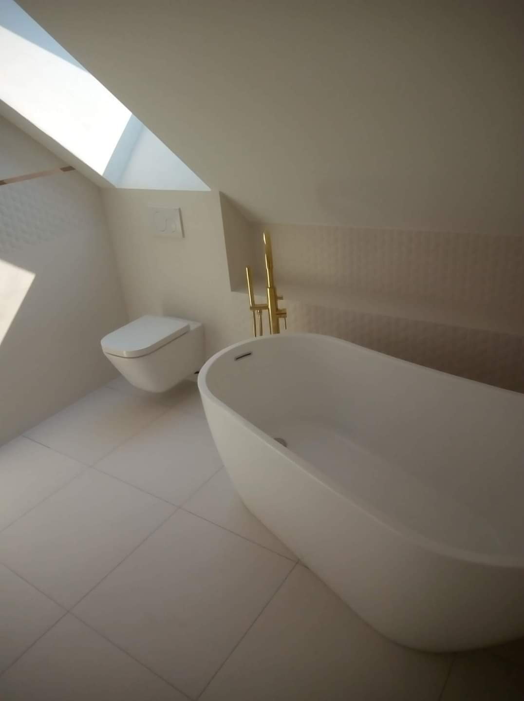 Nowoczesna łazienka na poddaszu z białą, wolnostojącą wanną i podwieszaną toaletą, złota armatura oraz okno dachowe wpuszczające naturalne światło.