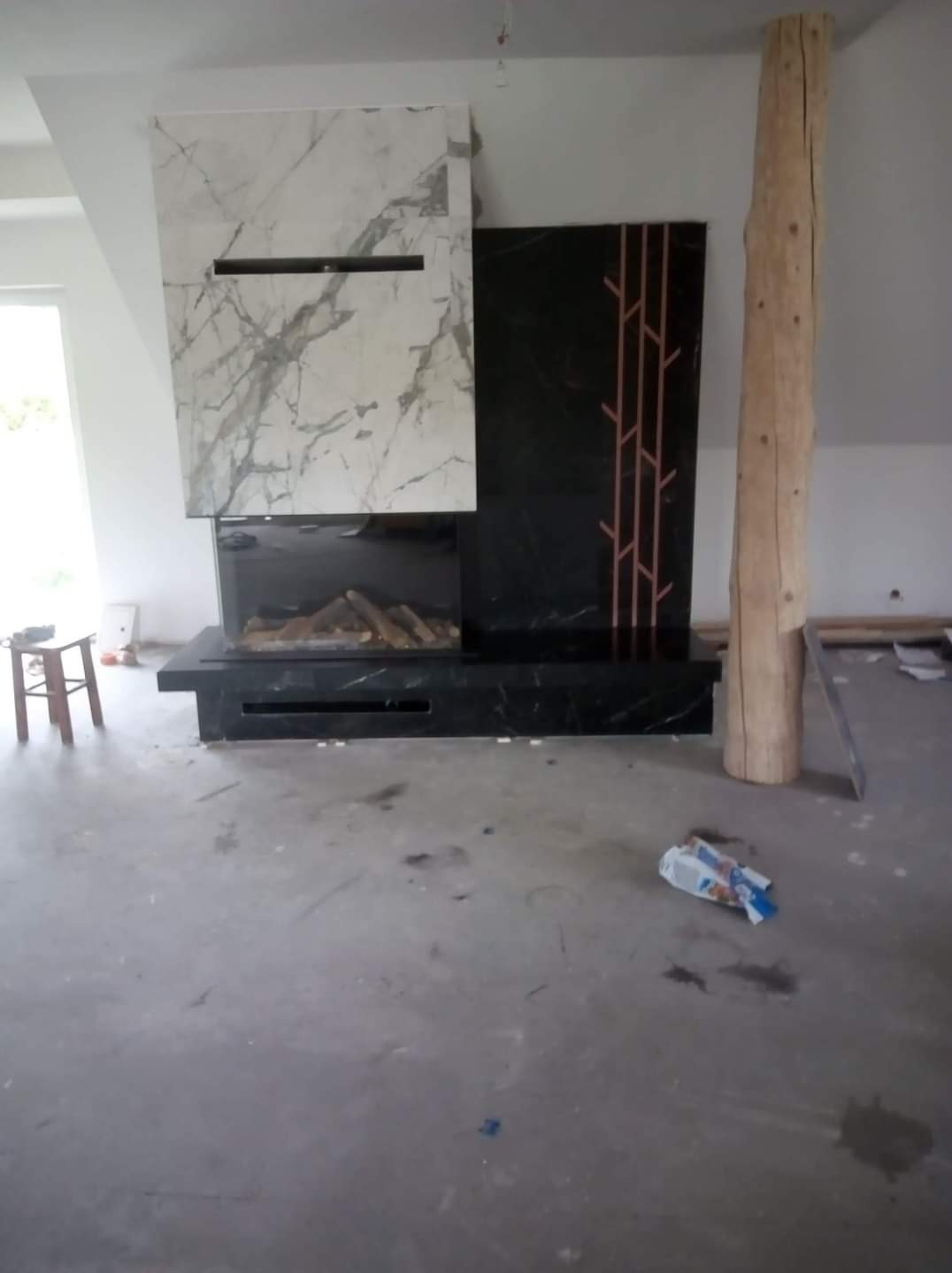 Nowoczesny kominek z marmurową obudową i dekoracyjnym panelem z motywem drzewa, surowy pień drzewa jako element wystroju, podłoga w trakcie remontu.