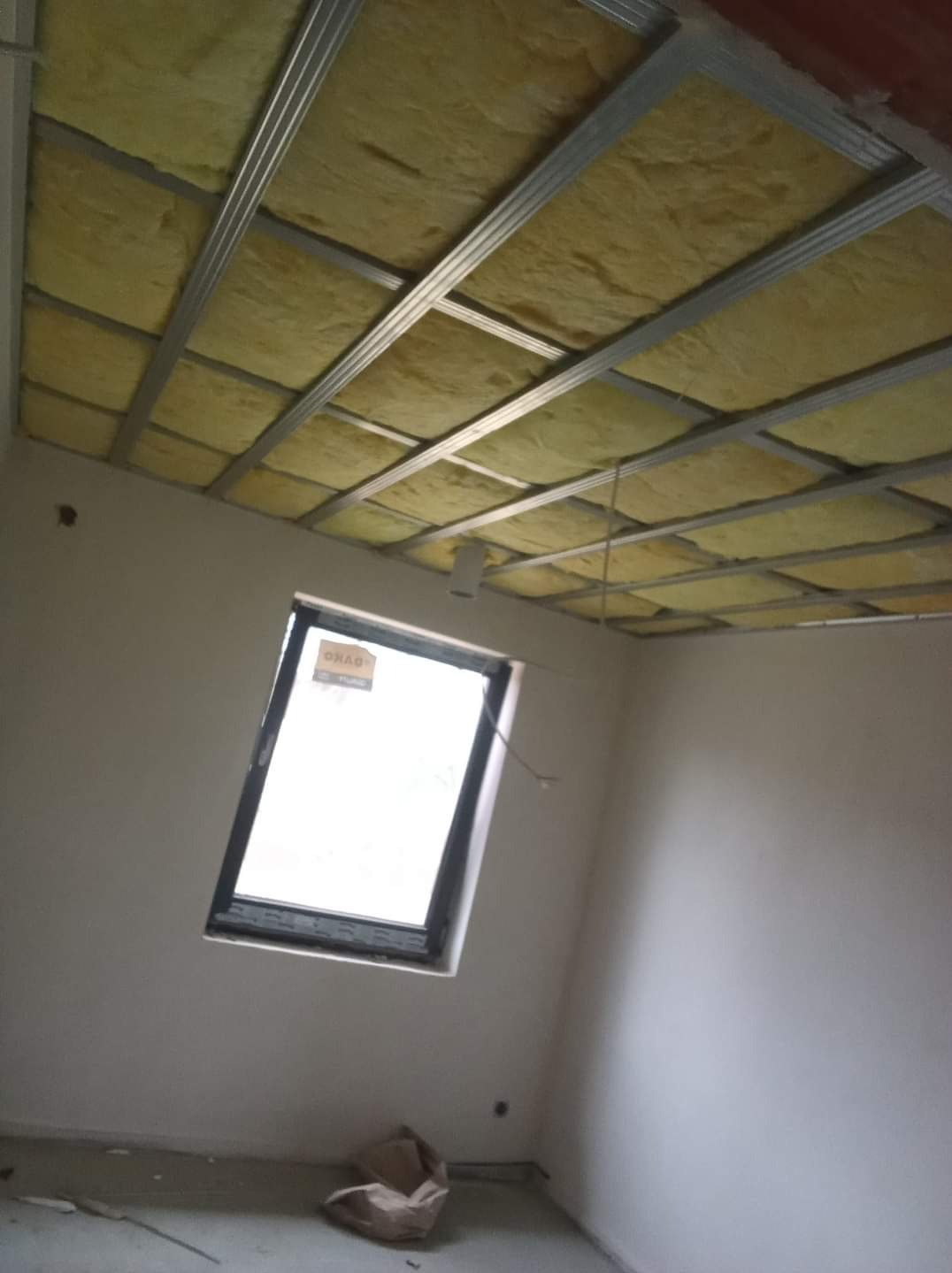 Montaż sufitu podwieszanego z izolacją z wełny mineralnej, widoczna konstrukcja metalowa i okno dachowe w surowym wnętrzu.