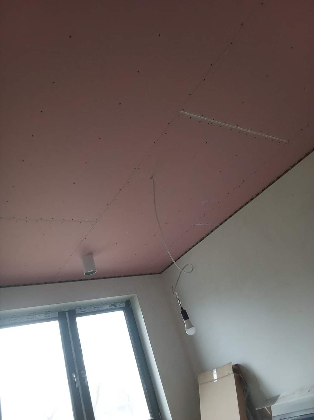 Różowa płyta karton gips na suficie z widocznymi śrubami, lampa sufitowa i prowizoryczne oświetlenie na kablu, okno w tle.