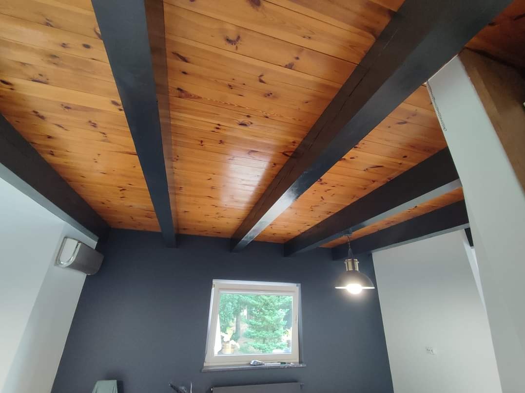 Drewniany sufit z belkami pomalowanymi na ciemny kolor, szara ściana, okno z widokiem na zieleń i lampa wisząca w stylu industrialnym.