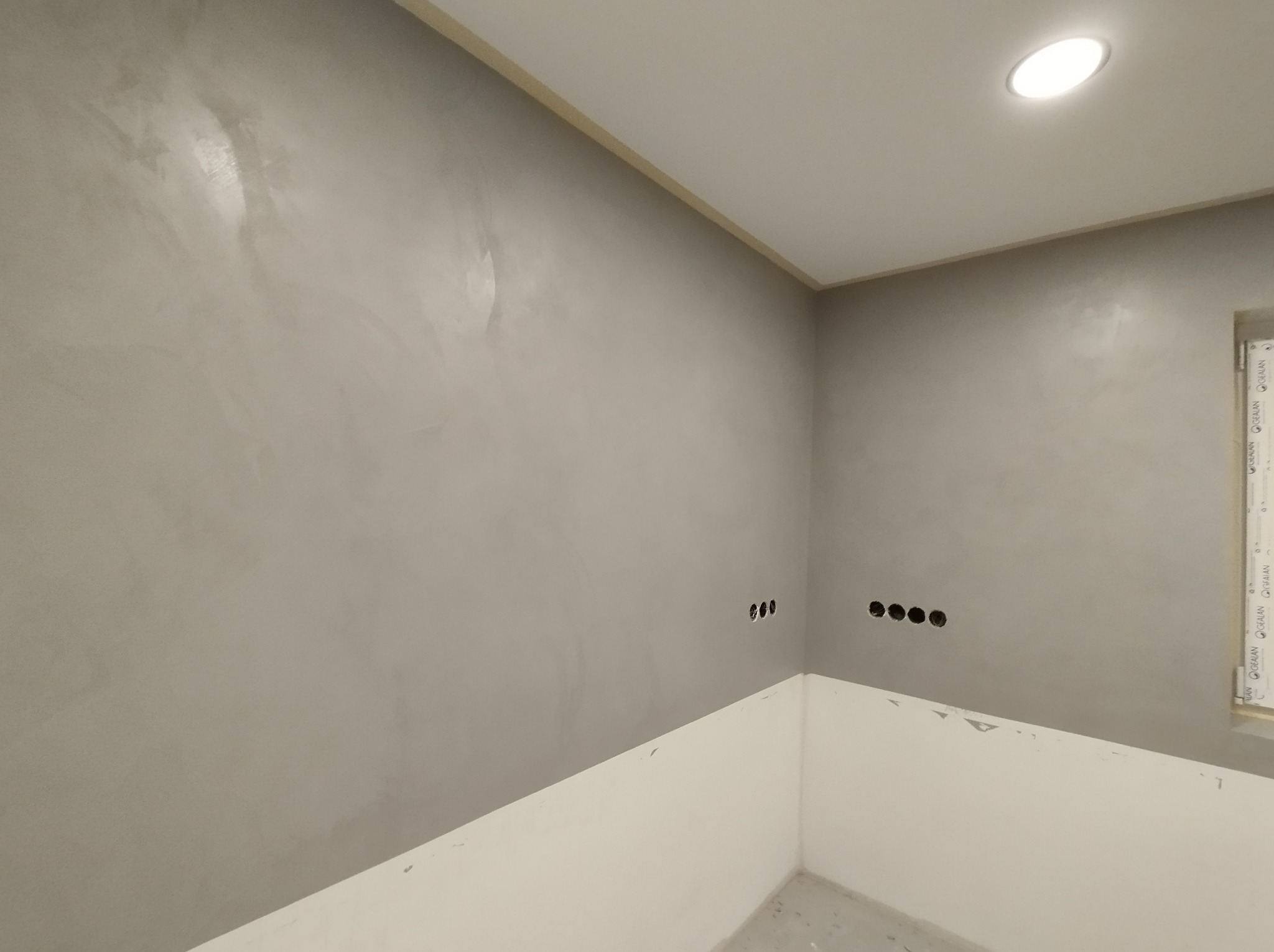 Ściana w odcieniach szarości z efektem dekoracyjnym, widoczne otwory na gniazdka elektryczne i biała listwa przypodłogowa w nowo wykończonym wnętrzu.