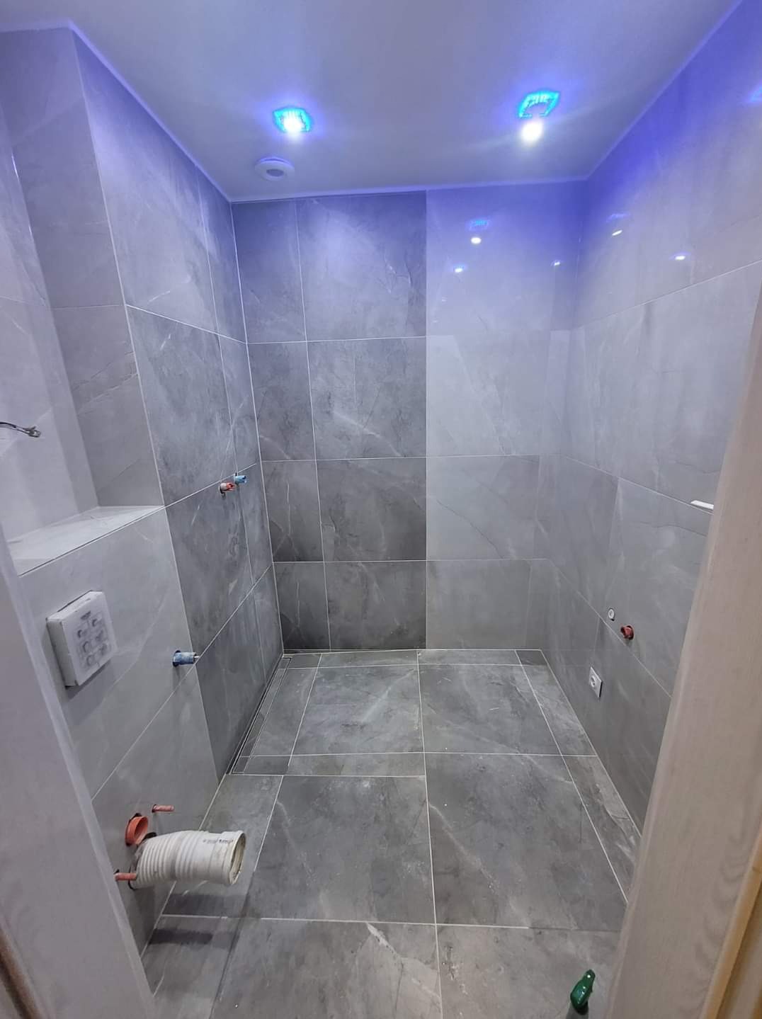 Wnętrze łazienki wykończone szarymi płytkami imitującymi marmur, z niebieskim oświetleniem LED na suficie, widoczne przyłącza wodne i odpływ liniowy w podłodze.