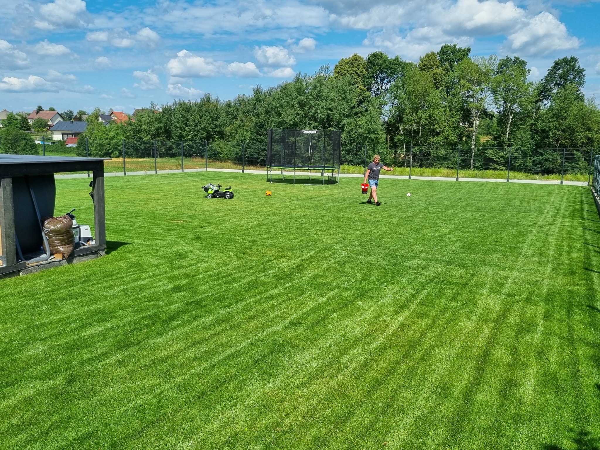 Rozległy trawnik z widocznymi pasami po koszeniu, na którym stoi trampolina, zabawkowy traktor i bawi się dziecko. W tle drzewa i domy.