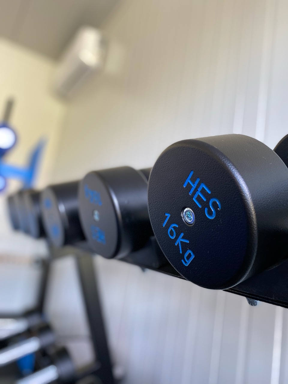 Rząd czarnych hantli 16kg z niebieskimi napisami HES na metalowym stojaku w siłowni, z klimatyzatorem widocznym w tle.