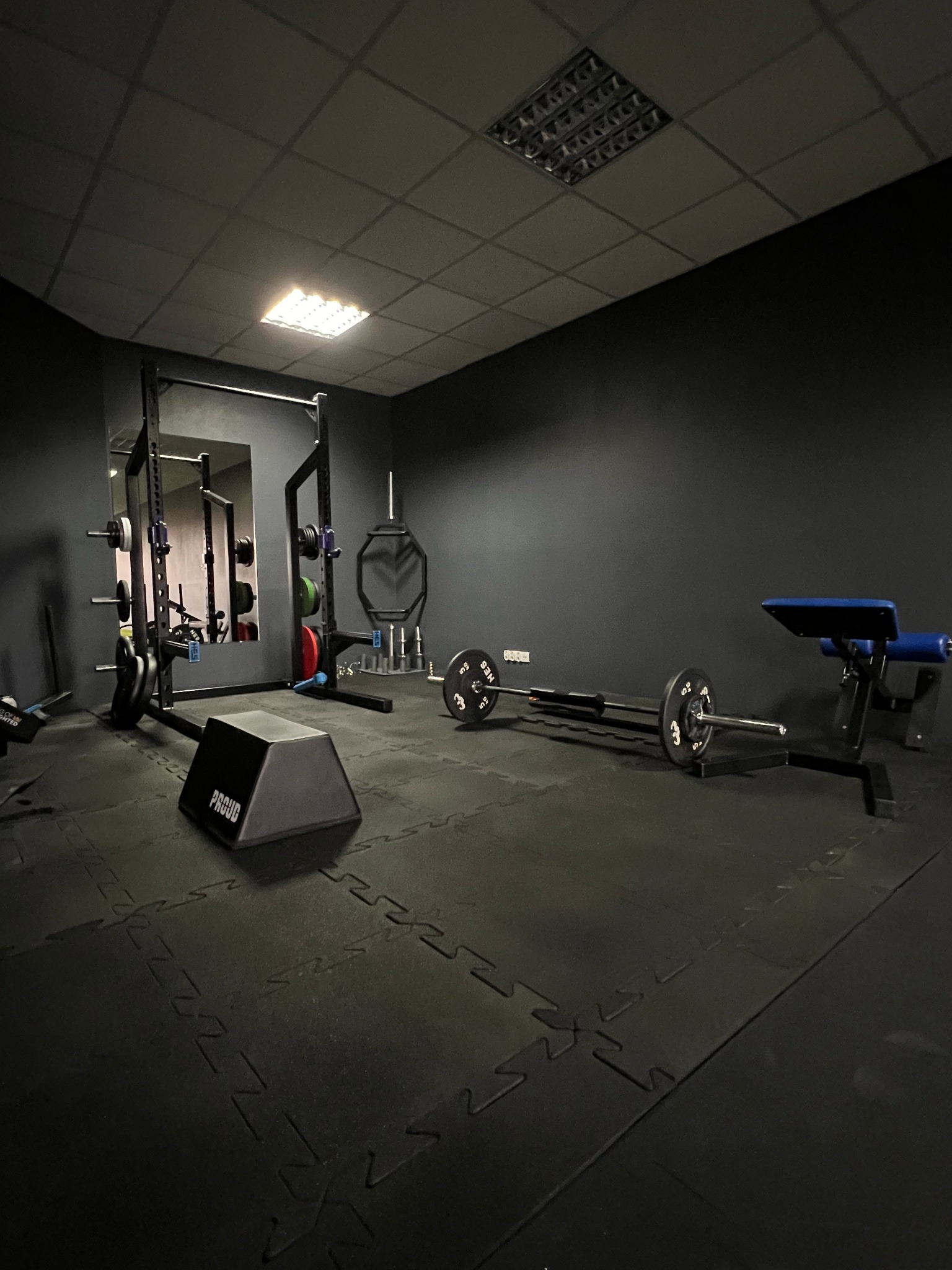 Nowoczesna siłownia z czarnymi ścianami, wyposażona w sprzęt do ćwiczeń siłowych: sztangi, stojaki, ławkę. Podłoga wyłożona matą. Minimalistyczny design.