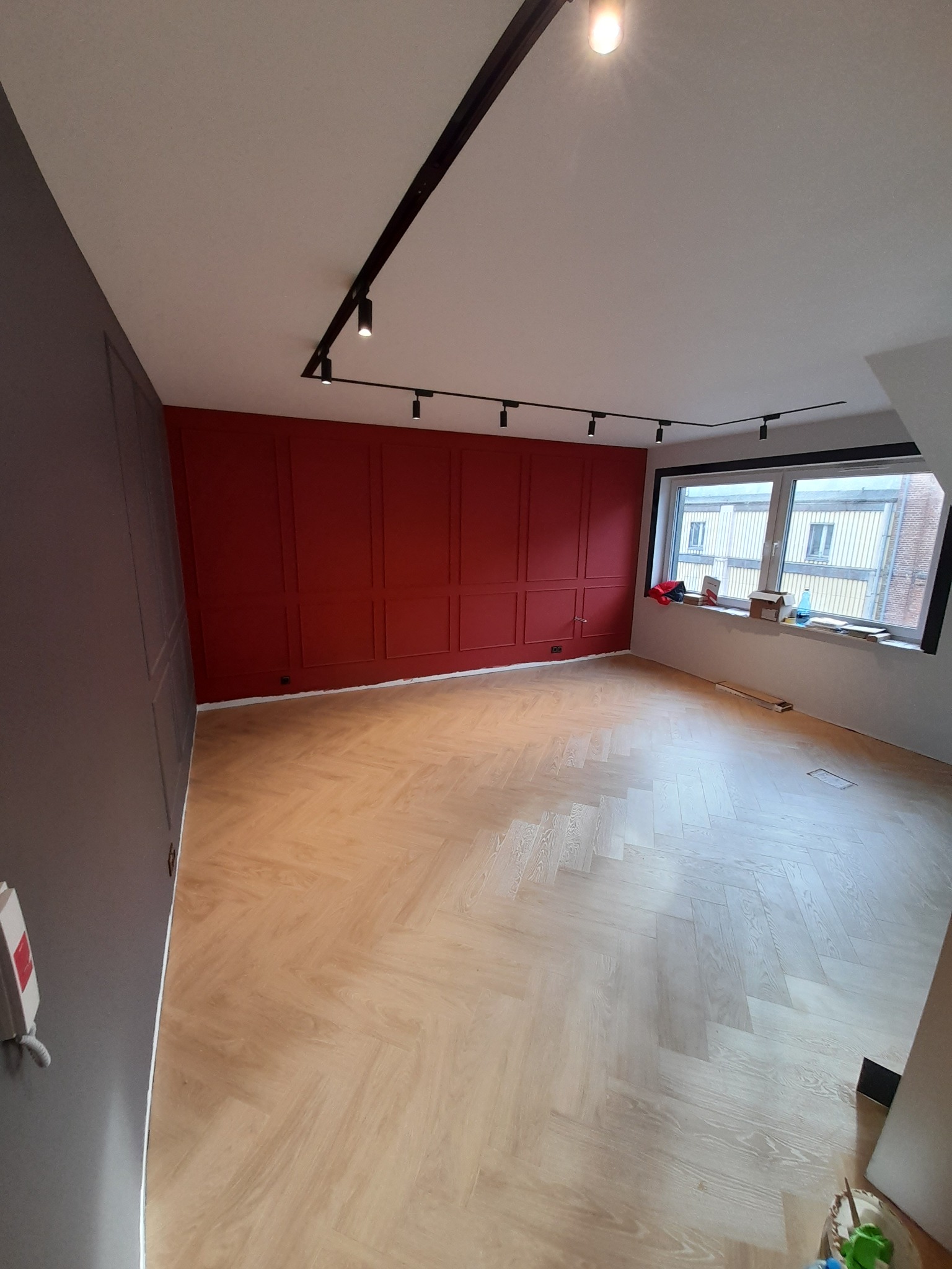 Jasna, drewniana podłoga ułożona w jodełkę w pokoju z czerwono-szarą ścianą i nowoczesnym oświetleniem szynowym.