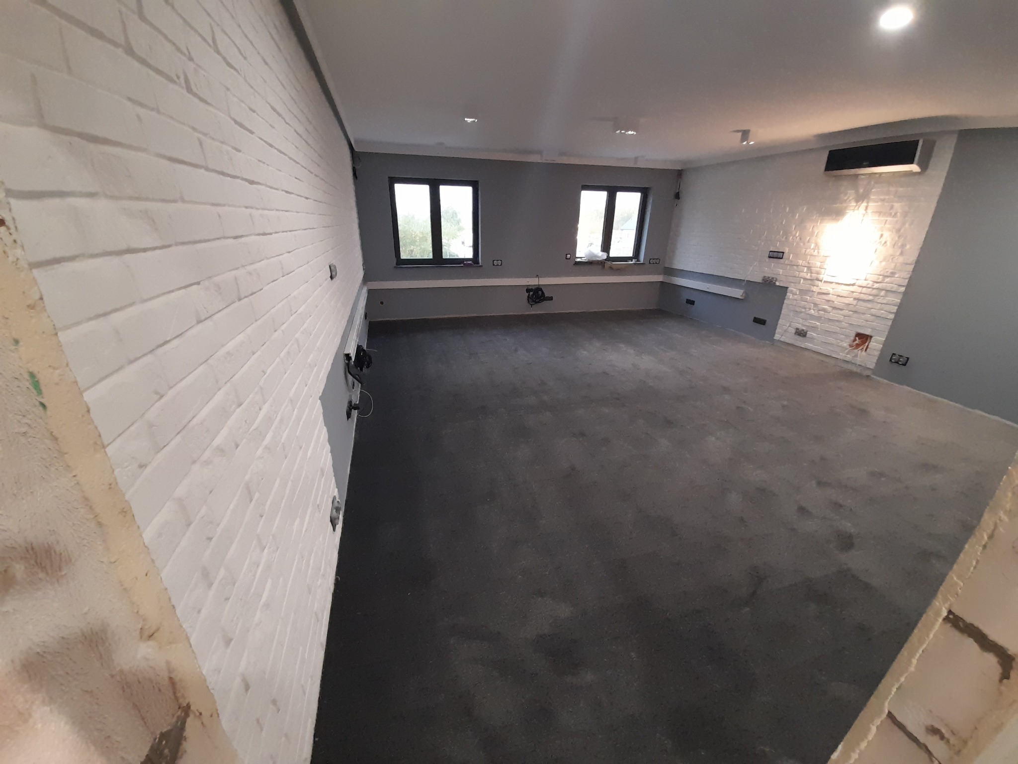 Surowe wnętrze biurowe z szarą wykładziną dywanową, białą cegłą na ścianach i dwoma oknami, w trakcie prac wykończeniowych, widoczne punkty elektryczne.