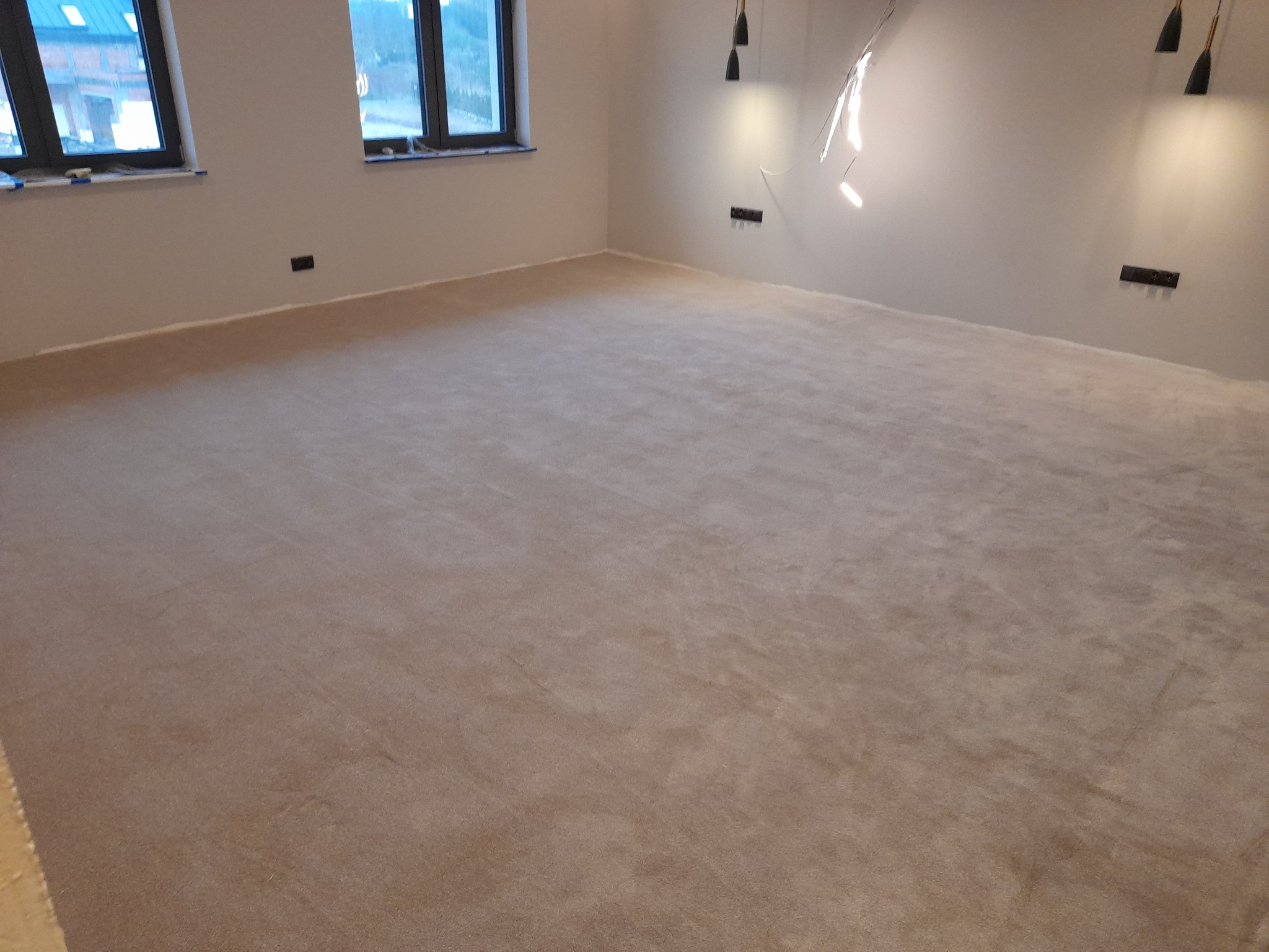 Beżowa, jednolita wykładzina dywanowa w pustym pokoju z dwoma oknami i nowoczesnym oświetleniem sufitowym.