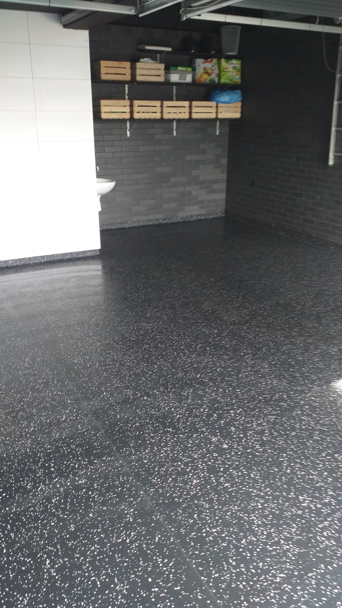 Błyszcząca, ciemnoszara podłoga z drobnymi, jasnymi plamkami w garażu z szarą cegłą i białymi płytkami na ścianach. Na półce drewniane skrzynki.