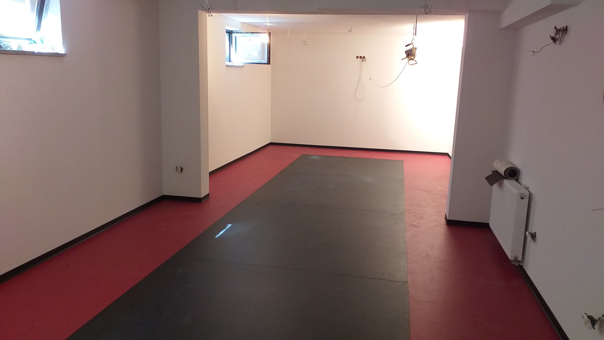 Przygotowanie do prac wykończeniowych w pomieszczeniu z czerwoną podłogą, widoczne płyty ochronne na podłodze, ściany przygotowane do malowania, grzejnik z rolką folii malarskiej.