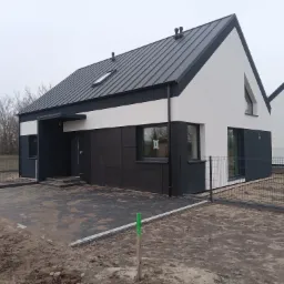 Nowoczesny dom jednorodzinny z czarno-białą elewacją i ciemnym dachem z blachy na tle pochmurnego nieba, widoczny fragment ogrodzenia i utwardzony podjazd z kostki brukowej.