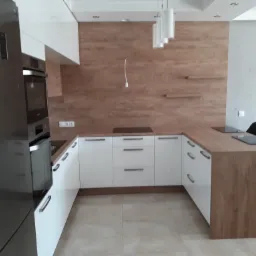 Nowoczesna kuchnia z białymi szafkami i blatem w kolorze drewna, zintegrowana płyta indukcyjna, stalowa lodówka i oświetlenie punktowe.