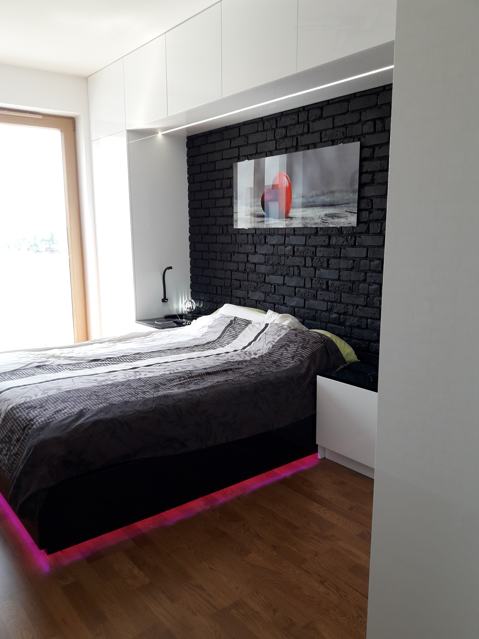 Nowoczesna sypialnia z łóżkiem, czarną ścianą z cegły i białymi szafami w zabudowie, oświetlenie LED pod łóżkiem, obraz na ścianie.