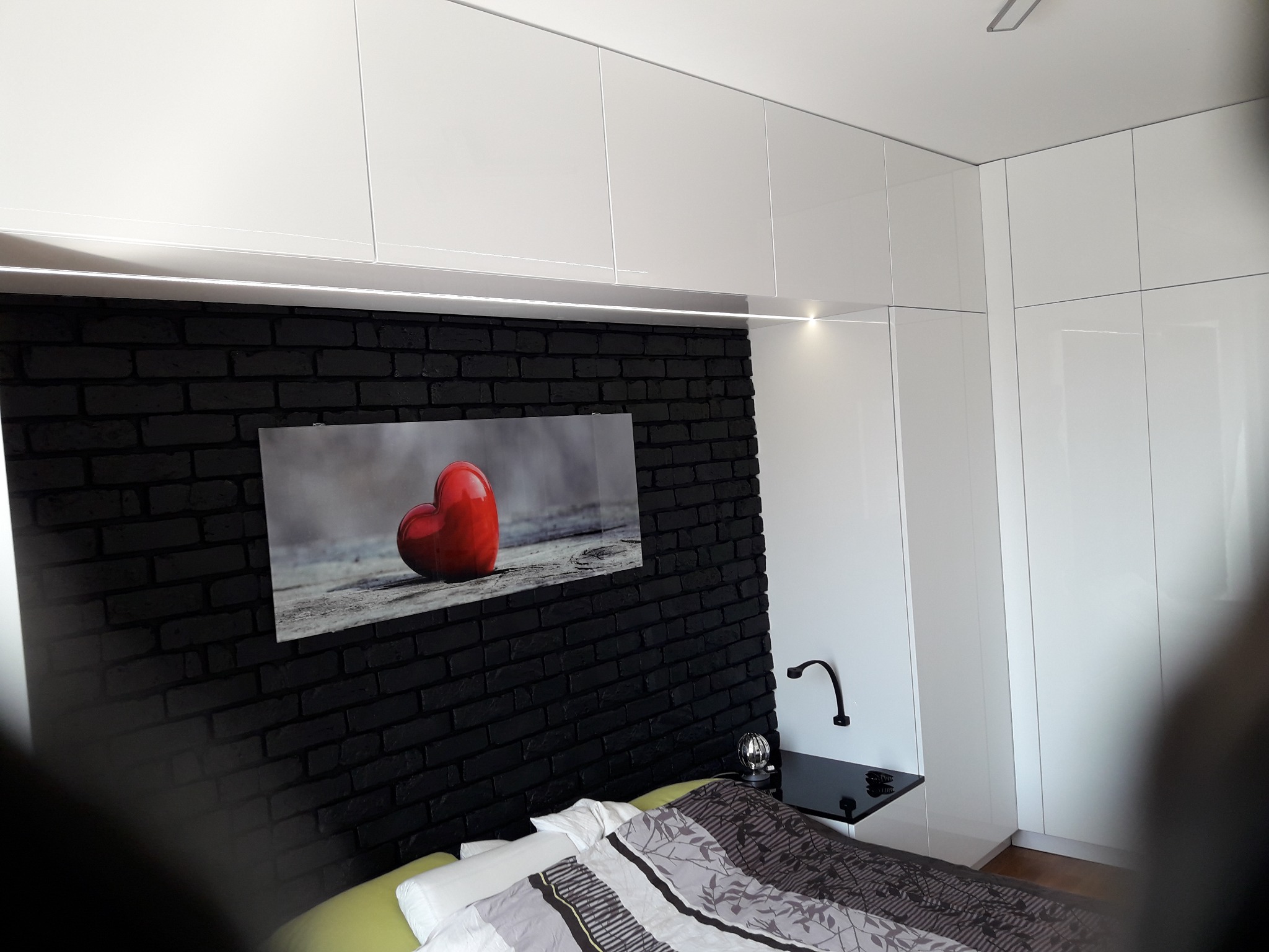 Szafa wnękowa z białymi, lakierowanymi frontami, oświetleniem LED podszafkowym i czarną ścianą z cegły, nad łóżkiem z pościelą w odcieniach szarości i czerni.