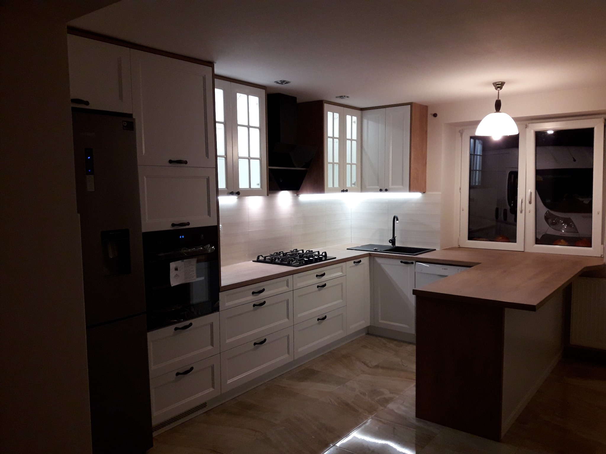 Nowoczesna kuchnia z białymi szafkami z czarnymi uchwytami, drewnianym blatem i wyspą, oświetleniem podszafkowym LED i marmurową podłogą.