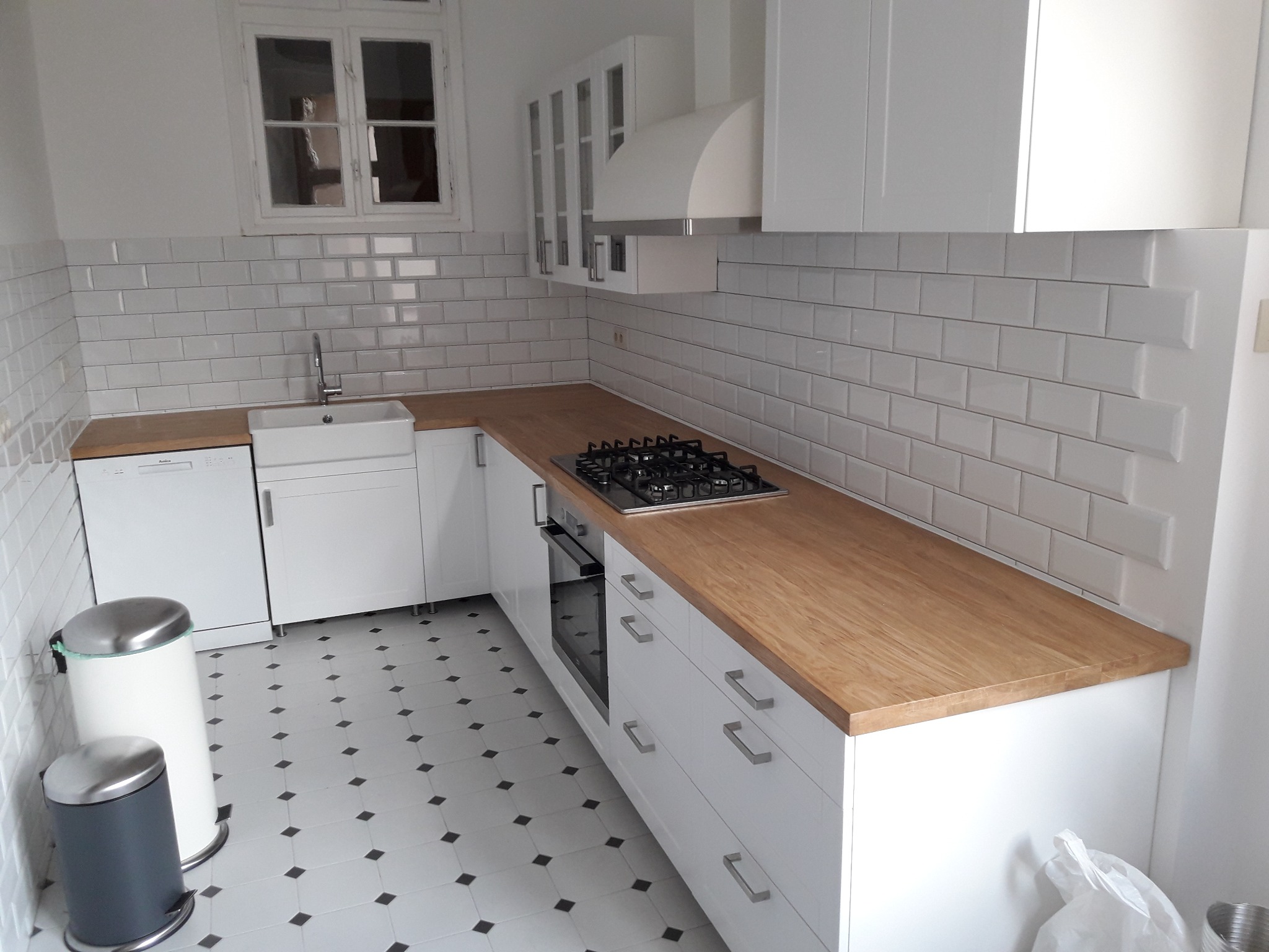 Jasna kuchnia z białymi szafkami, drewnianym blatem i białymi płytkami na ścianie, widok na zlew, kuchenkę gazową i kosze na śmieci.