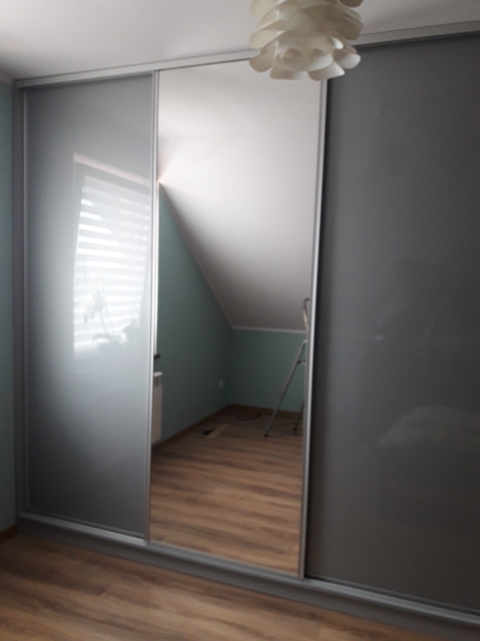 Szafa wnękowa z przesuwnymi drzwiami: szare matowe panele boczne i lustrzany panel środkowy odbijający skos dachu i drabinę w pokoju na poddaszu.