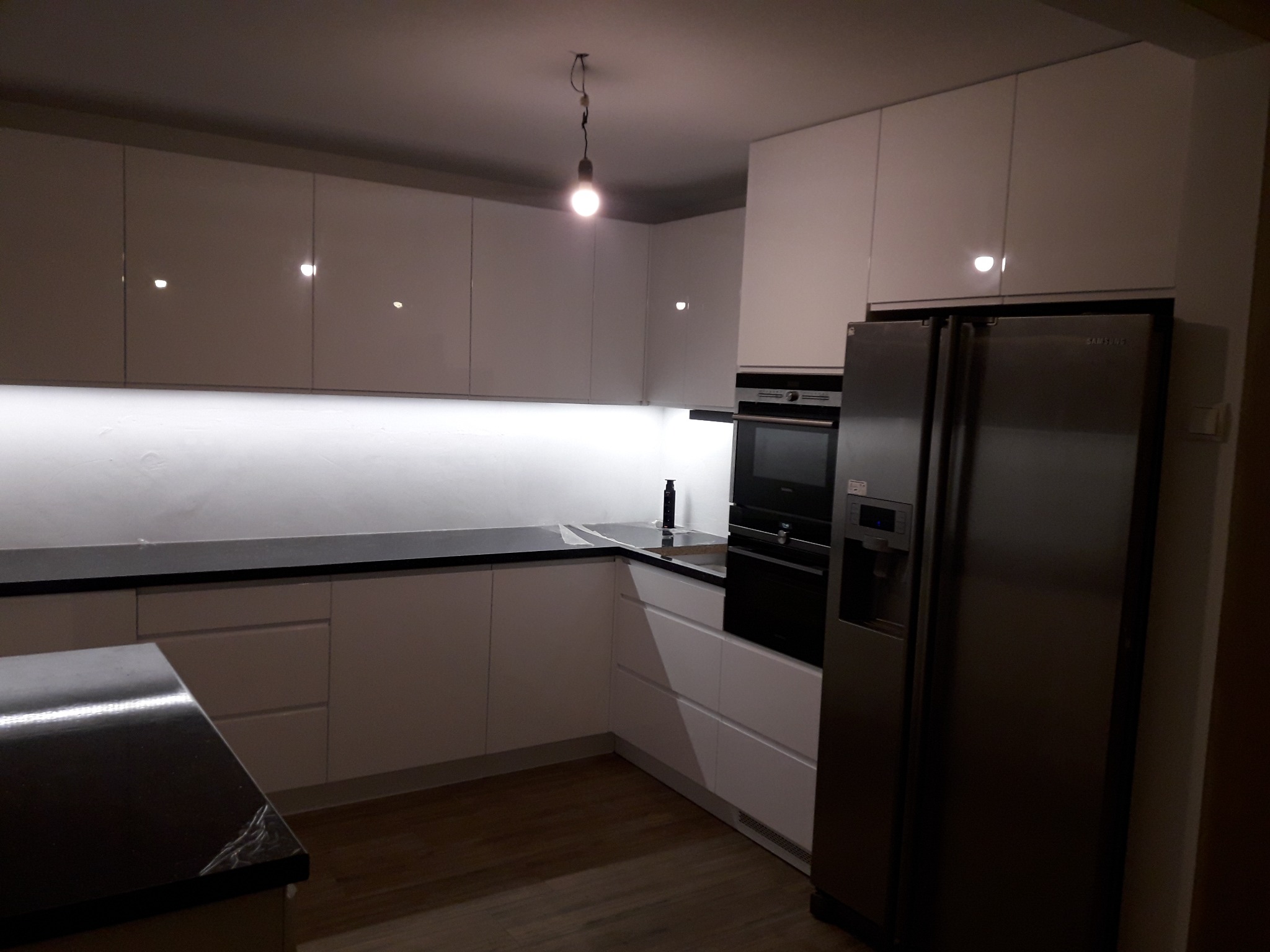Nowoczesna biała kuchnia z połyskiem, czarny blat, oświetlenie LED pod szafkami, lodówka Samsung. Widoczny fragment blatu z folią ochronną.
