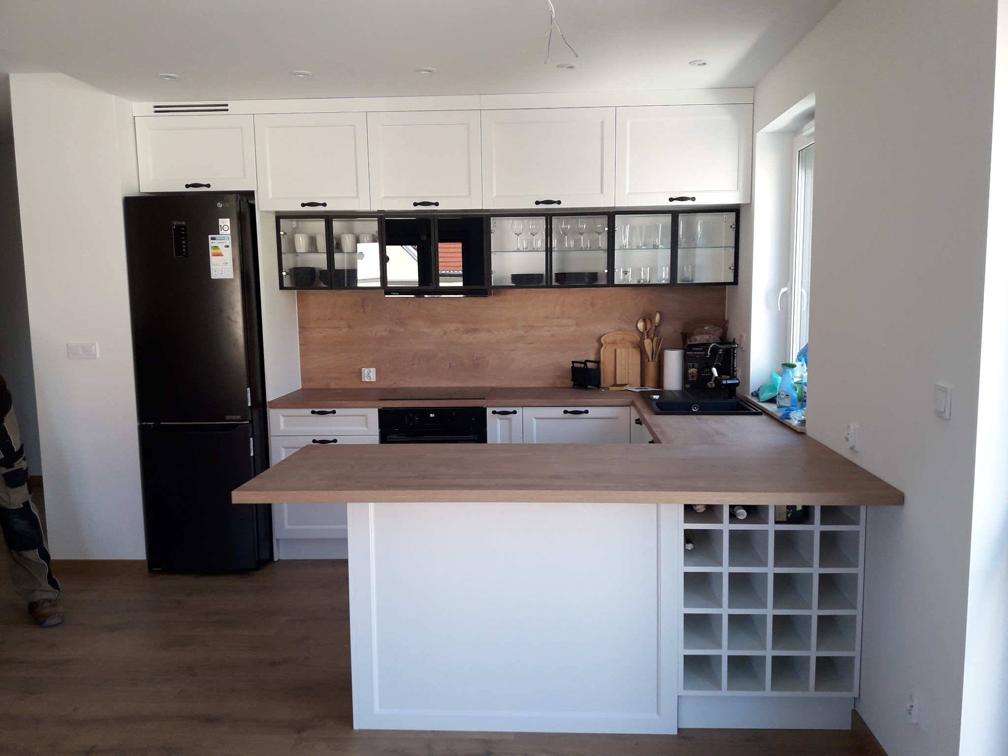 Jasna kuchnia z drewnianym blatem w Dziadowej Kłodzie, z wyspą i miejscem na wino, czarna lodówka obok białych szafek.