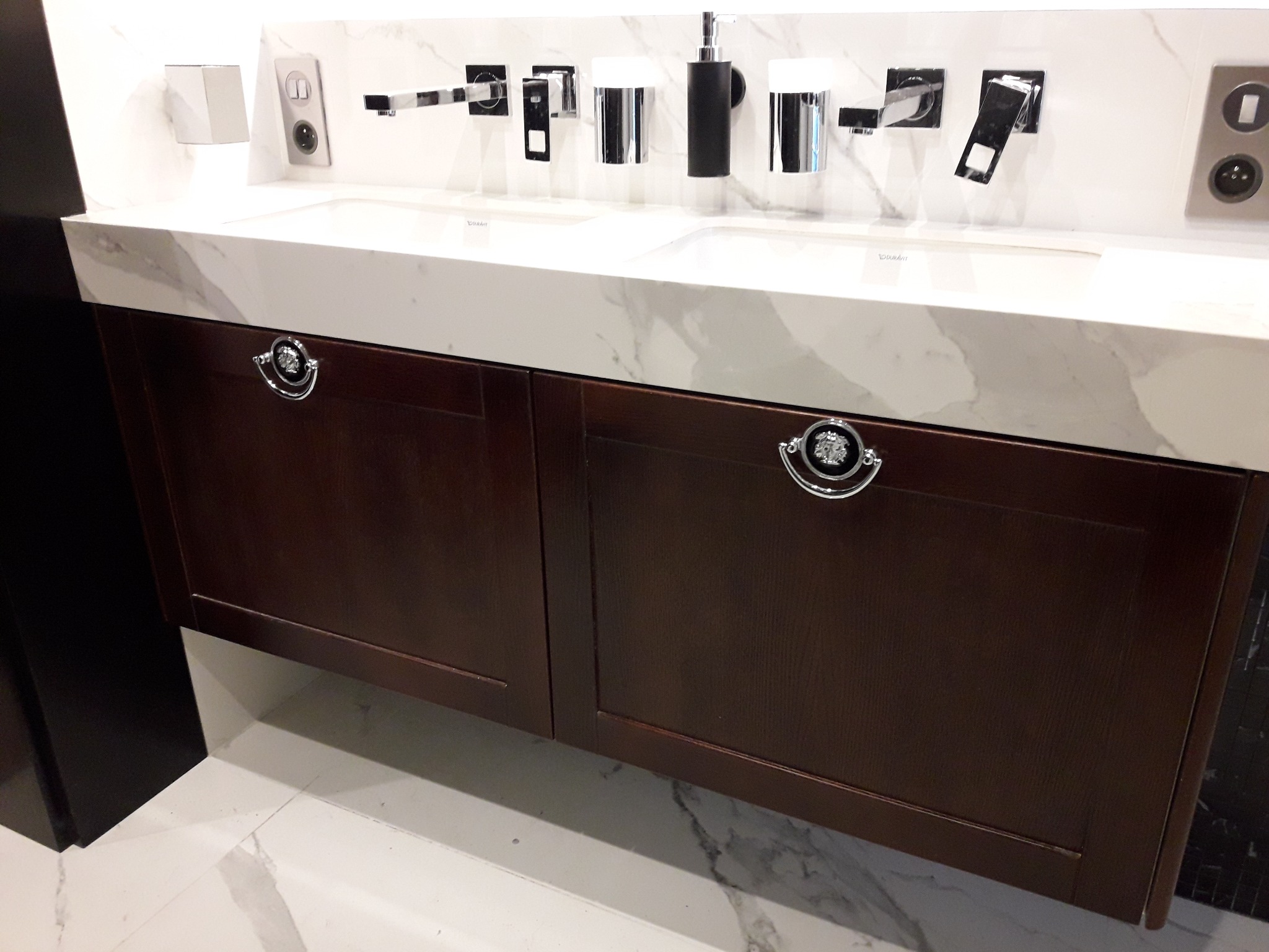 Dwukomorowa umywalka z białym blatem imitującym marmur i brązową szafką z ozdobnymi uchwytami, armatura ścienna w nowoczesnym stylu.