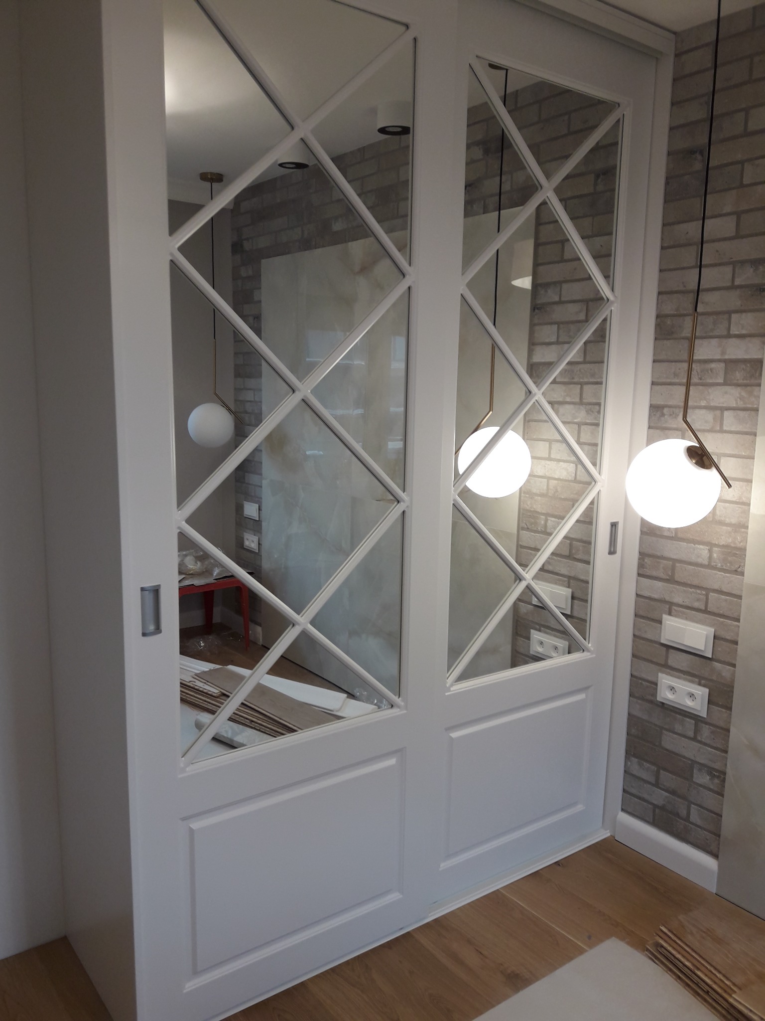 Biała szafa przesuwna z lustrzanymi drzwiami w kratkę, widoczne elementy wykończeniowe i wiszące lampy kuliste