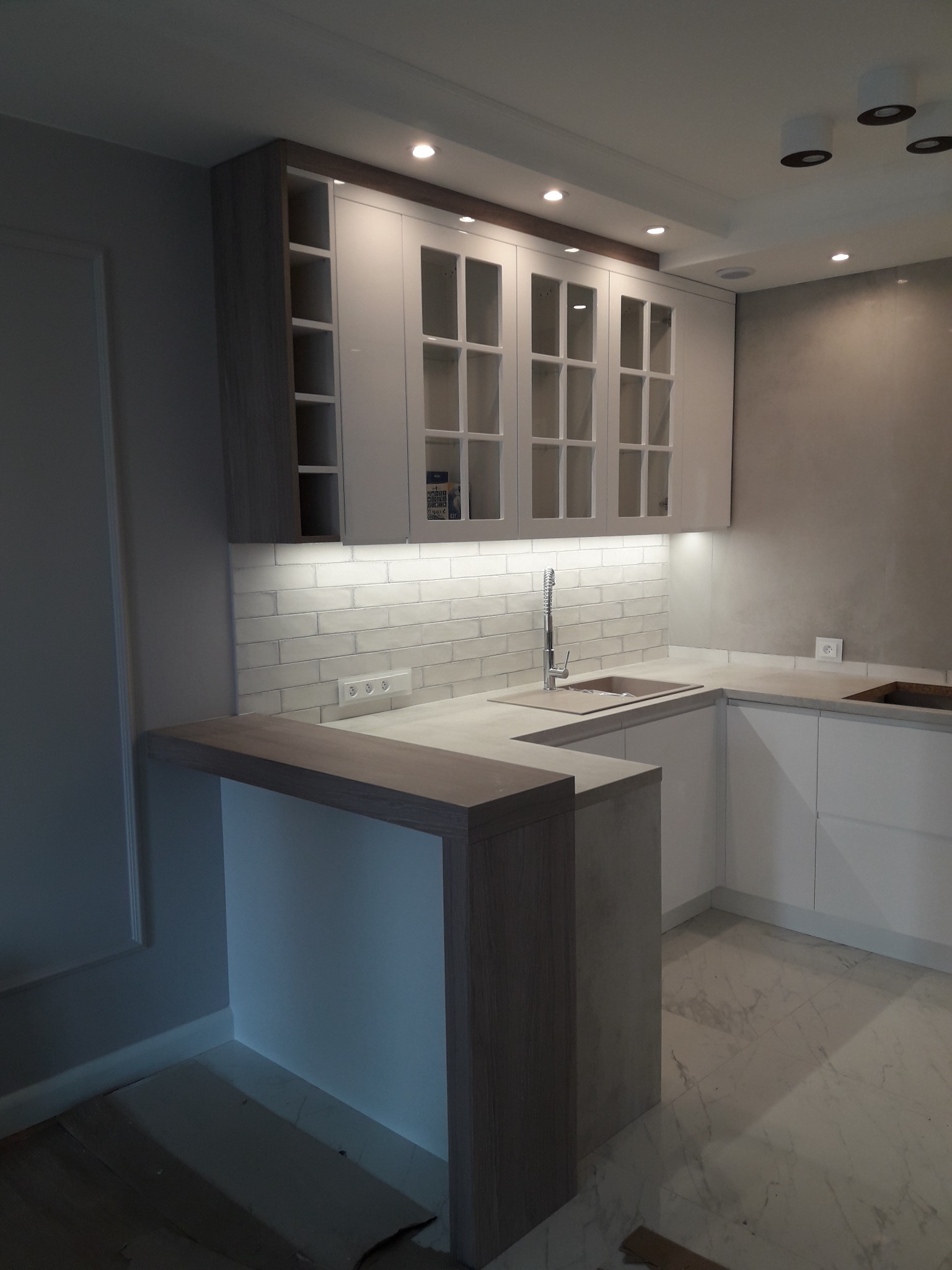 Nowoczesna kuchnia z białymi frontami szafek, drewnianymi akcentami blatu i barku, podświetleniem LED oraz marmurową podłogą.