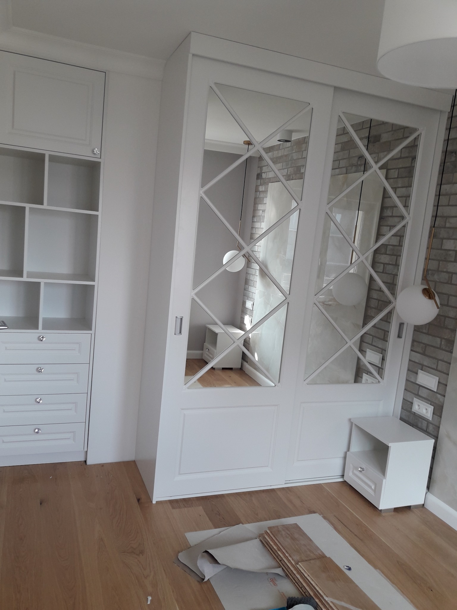 Biała szafa przesuwna z lustrami w romby, dopasowana do zabudowy z półkami i szufladami, drewniana podłoga w trakcie montażu, widoczne materiały wykończeniowe.