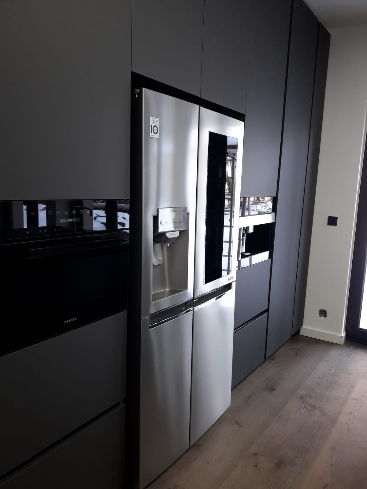Nowoczesna kuchnia z zabudową meblową w odcieniach szarości, zintegrowana z srebrną lodówką side-by-side i piekarnikiem marki Miele, na drewnianej podłodze.