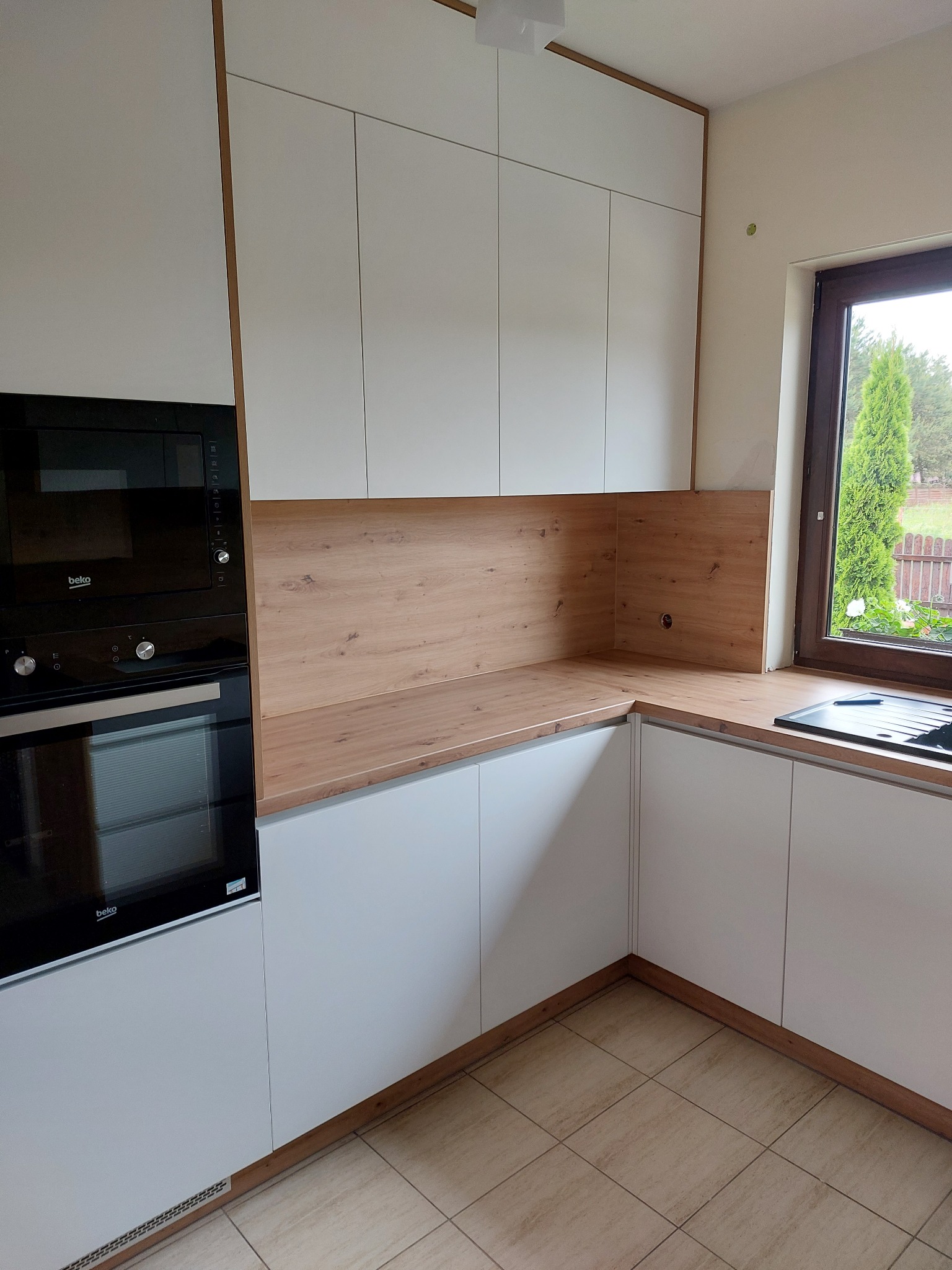 Nowoczesna kuchnia z białymi szafkami i drewnianym blatem, piekarnik w zabudowie, widok z perspektywy podłogi, okno z widokiem na ogród.