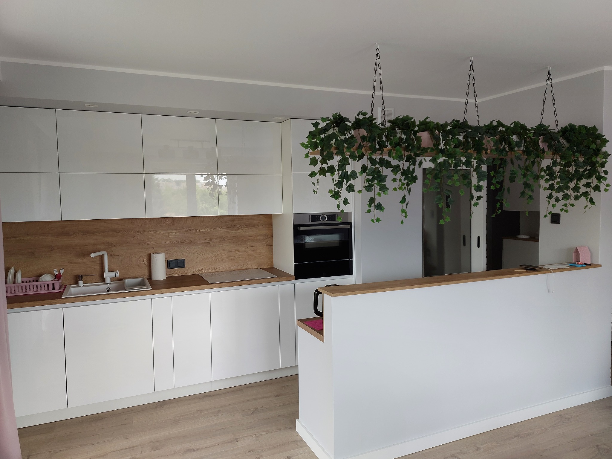 Nowoczesna kuchnia z białymi, lakierowanymi frontami, drewnianym blatem i wiszącymi roślinami doniczkowymi nad wyspą.