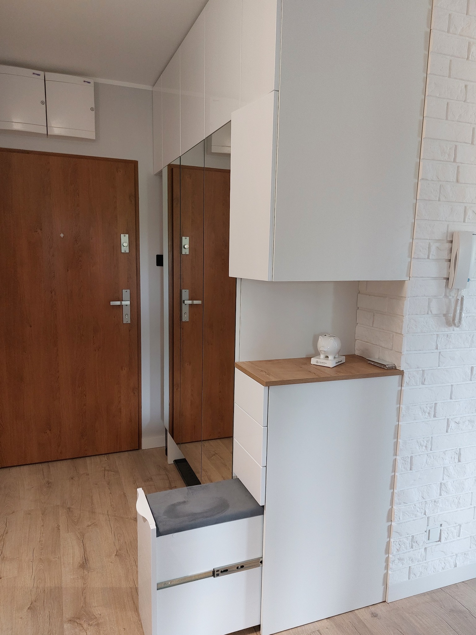 Biała szafa z siedziskiem i lustrem w przedpokoju, drewniane drzwi wejściowe i ściana z białej cegły w tle.