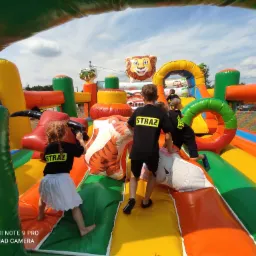 Dzieci ubrane w koszulki z napisem STRAŻ bawią się na kolorowym dmuchanym zamku z motywami zwierzęcymi, widok z perspektywy dziecka wewnątrz konstrukcji.