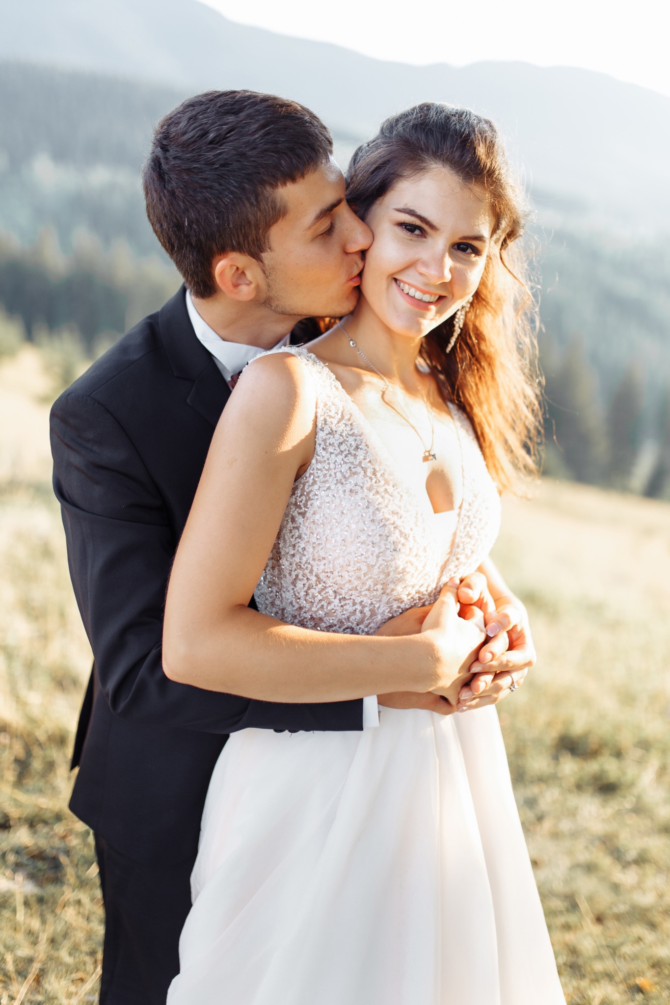Uśmiechnięta panna młoda w sukni z cekinami, obejmowana przez pana młodego w czarnym garniturze, który całuje ją w policzek, na tle górskiego krajobrazu w ciepłym świetle.
