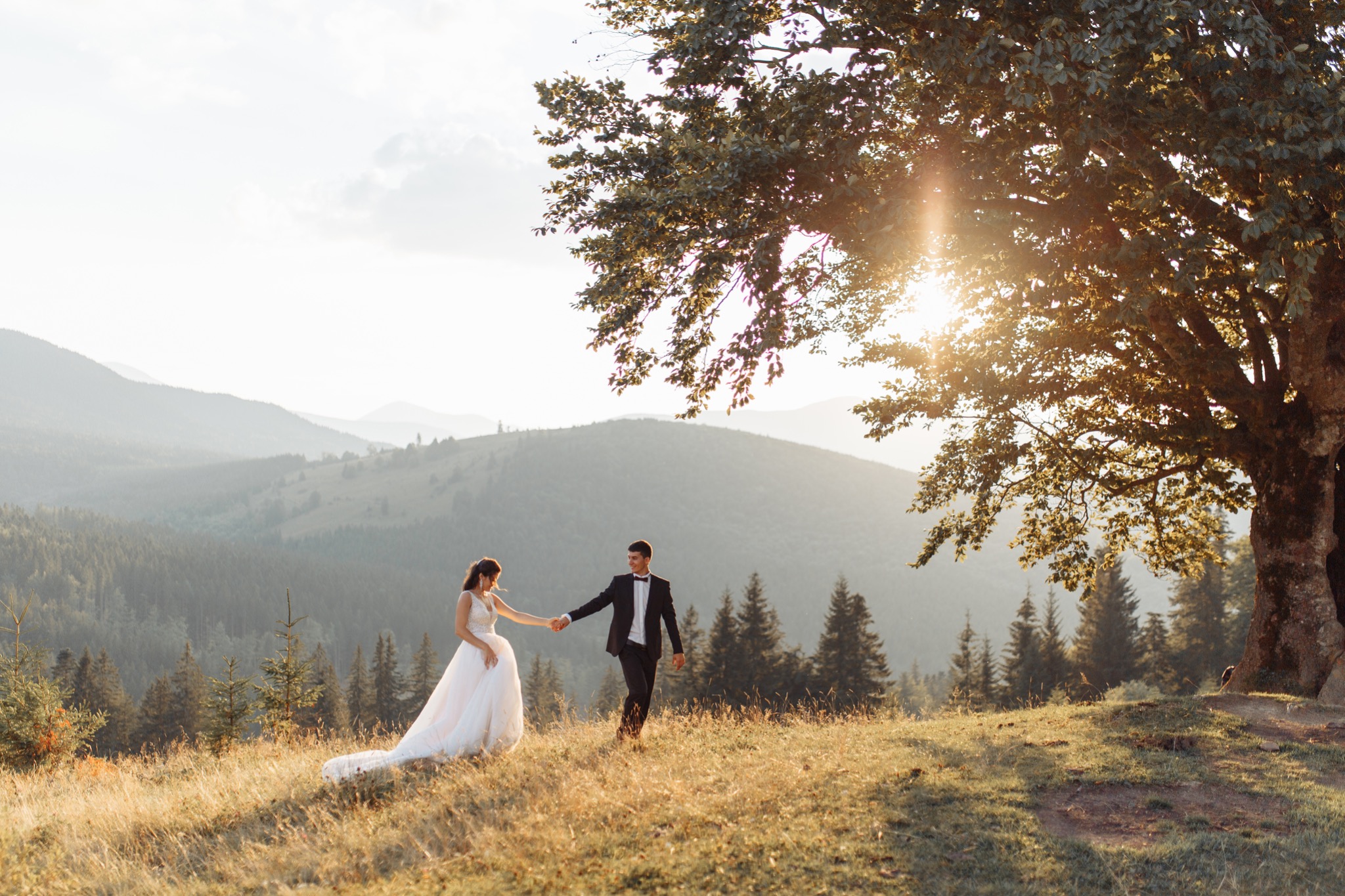 Para młoda trzymająca się za ręce spaceruje po wzgórzu w Łodzi, na tle gór i zachodzącego słońca, widok zza drzewa.