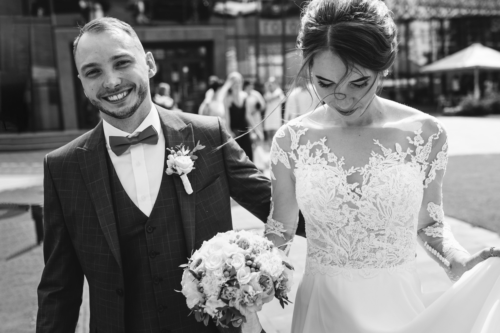 Czarno-białe zdjęcie uśmiechniętego pana młodego w kraciastej marynarce z bukietem oraz panny młodej w koronkowej sukni z opuszczonym wzrokiem, w tle rozmyte sylwetki ludzi.