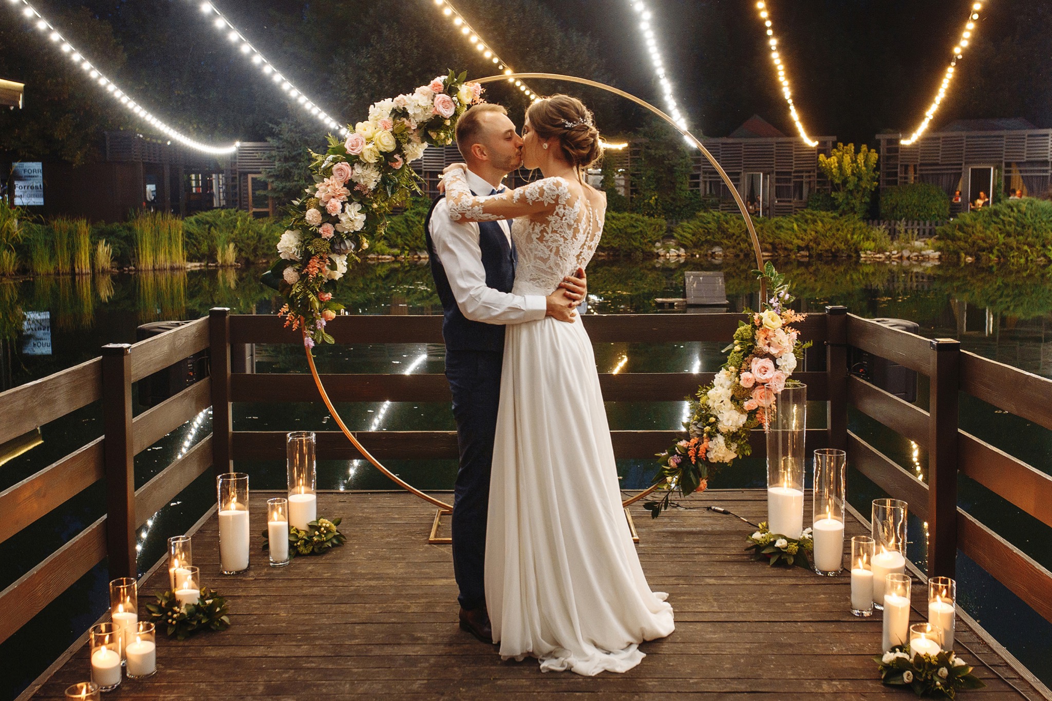 Pocałunek nowożeńców na drewnianym pomoście nad jeziorem, okrągła dekoracja kwiatowa, świece w szklanych wazonach, girlandy świetlne w tle.
