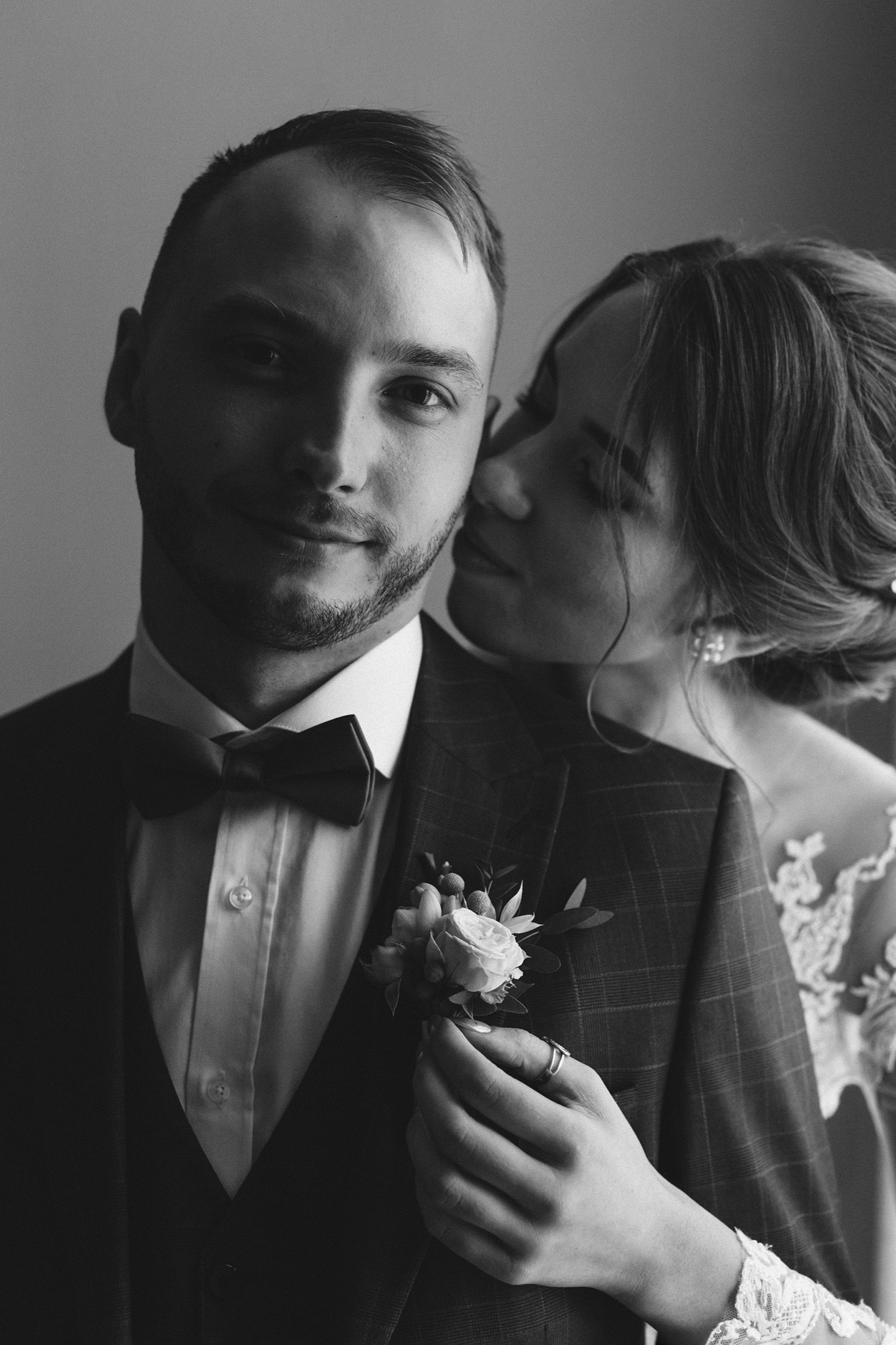 Czarno-białe zbliżenie pary młodej: uśmiechnięty pan młody w garniturze w kratę z muchą i butonierką, panna młoda obejmuje go, widać koronkę sukni i kolczyk.