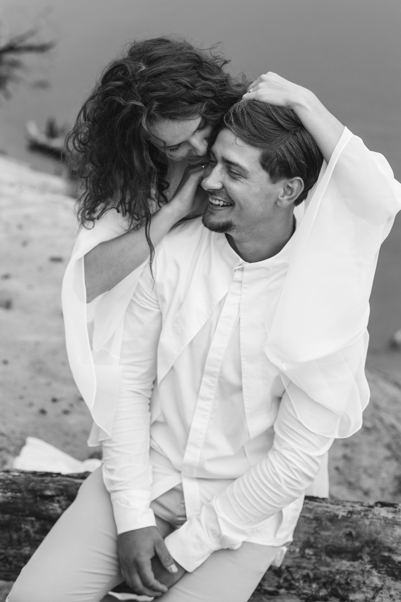Czarno-białe zdjęcie uśmiechniętej pary w jasnych ubraniach, kobieta obejmuje mężczyznę na tle plaży i wody.