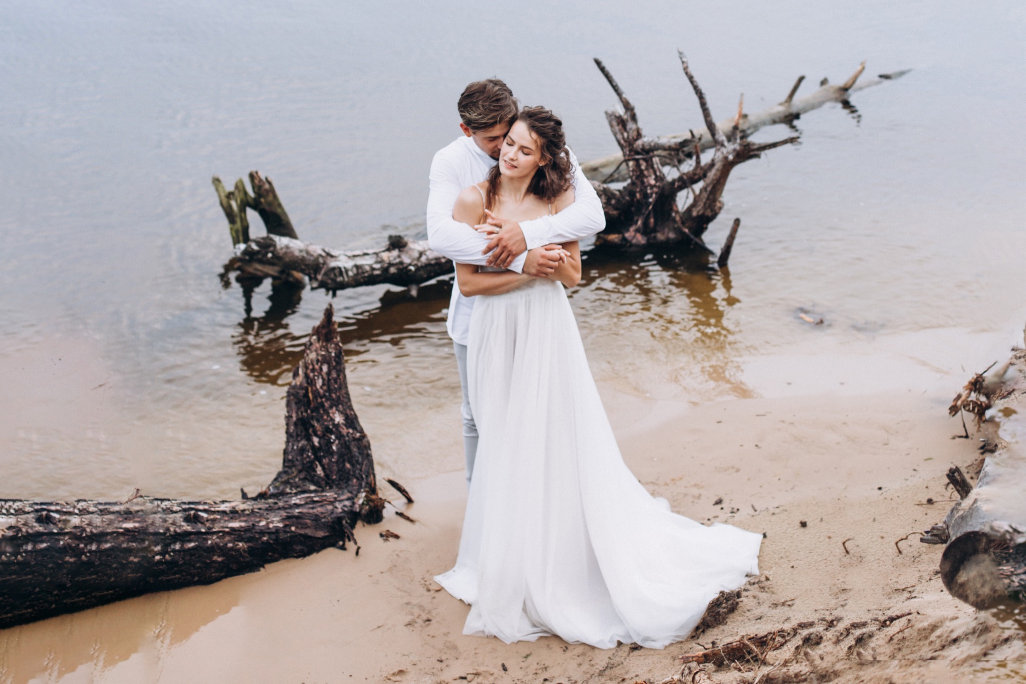 Para młoda w białych strojach, objęta na piaszczystej plaży z dryfującym drewnem, w tle woda.