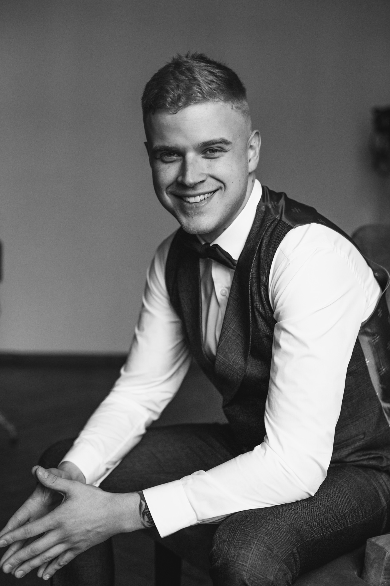 Portret uśmiechniętego mężczyzny w kamizelce i muszce, siedzącego na krześle, w czarno-białej tonacji, z widocznym tatuażem na nadgarstku.