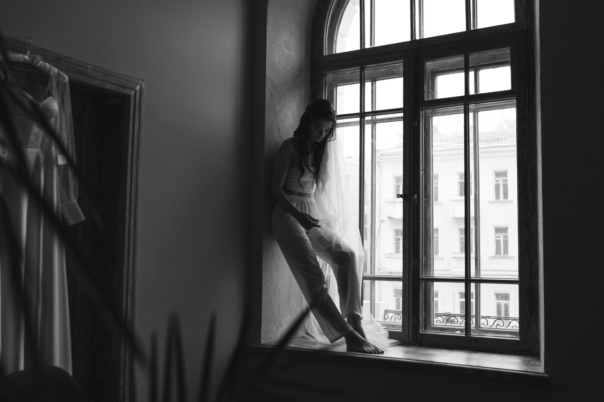 Czarno-białe zdjęcie panny młodej w bieliźnie, siedzącej na parapecie okna z widokiem na budynek, obok wisi suknia ślubna.