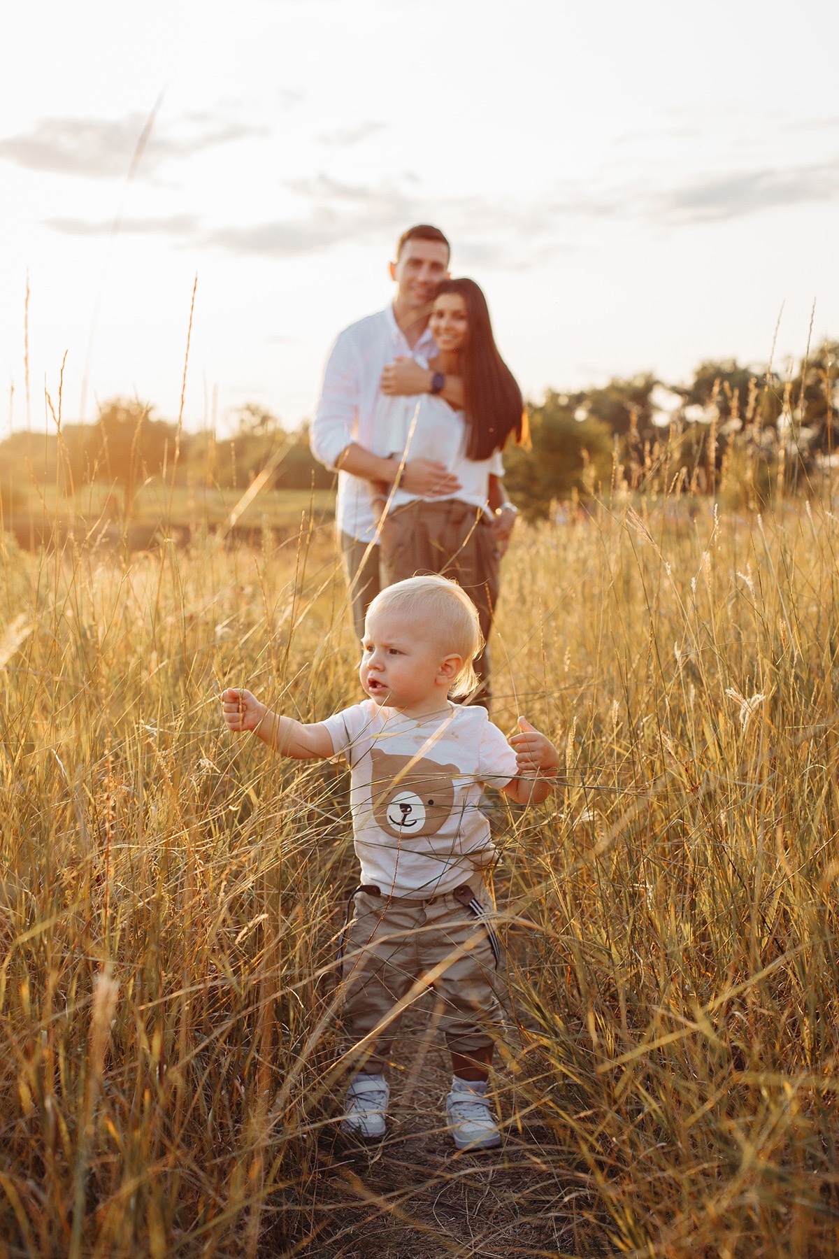 Mały chłopiec z blond włosami w koszulce z misiem idzie przez pole trawy, w tle uśmiechnięta para w objęciach.