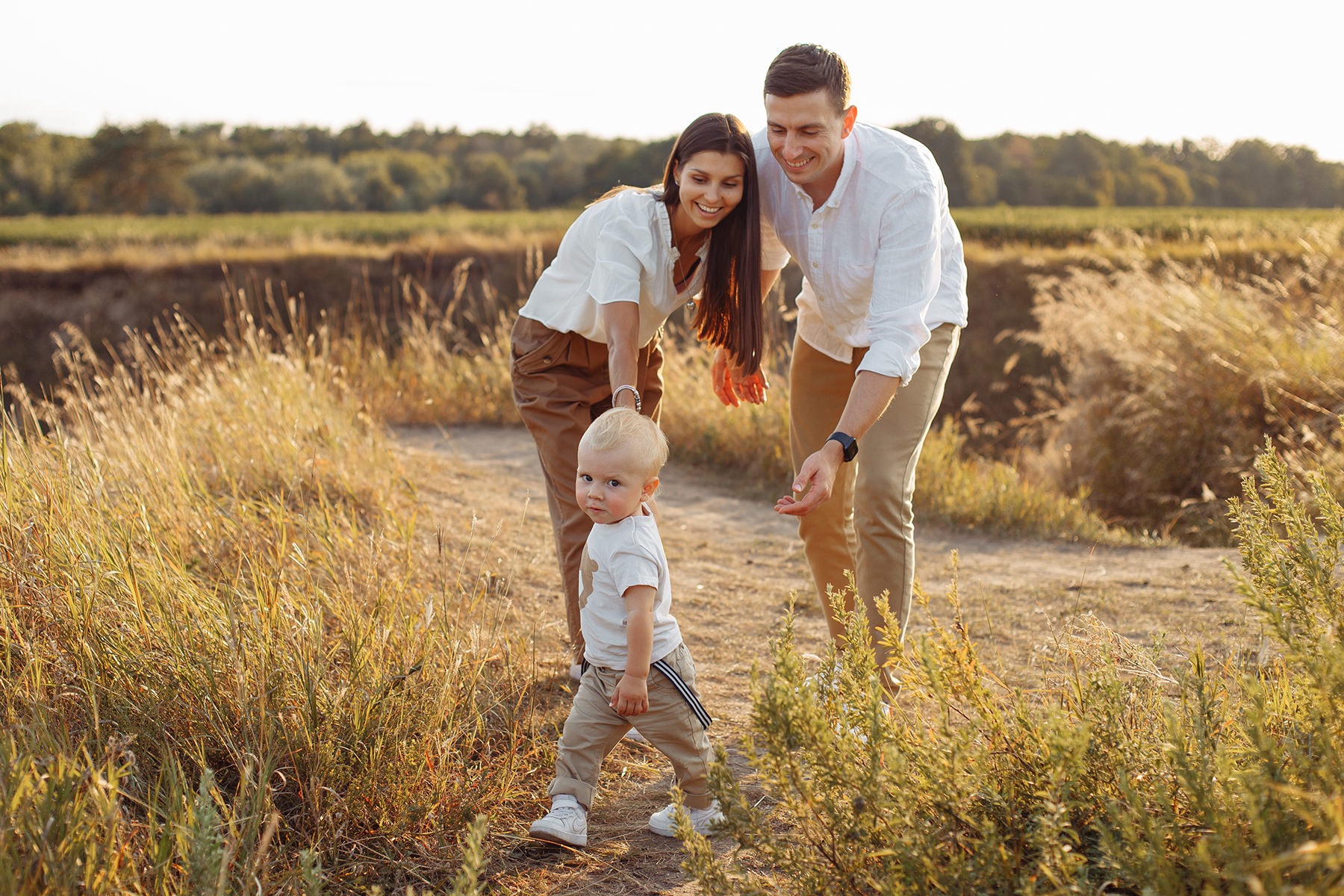 Rodzinne zdjęcie w plenerze: młoda para z blondwłosym synkiem, który stawia pierwsze kroki na polnej ścieżce w otoczeniu wysokich traw w ciepłym, zachodzącym słońcu.