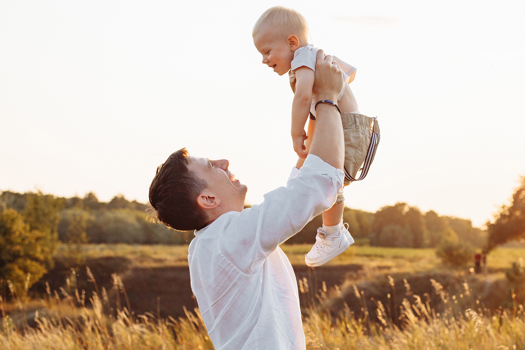 Uśmiechnięty tata w białej koszuli podnosi wysoko małego synka w beżowych spodenkach z szelkami na tle łąki w promieniach zachodzącego słońca.