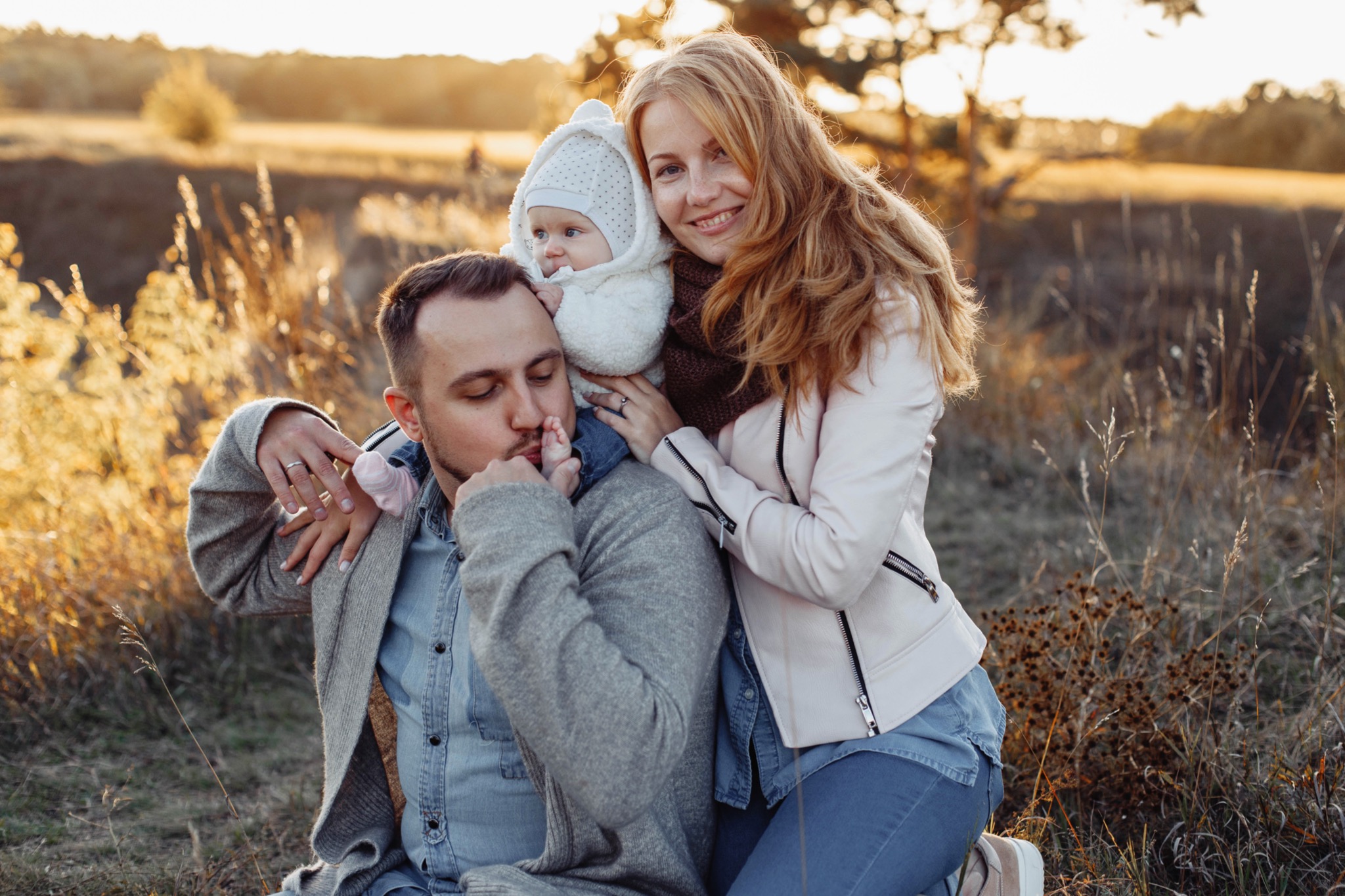 Rodzinne ujęcie w plenerze: tata całuje stópkę niemowlęcia w białym kombinezonie, mama z uśmiechem obejmuje dziecko na tle łąki o zachodzie słońca.