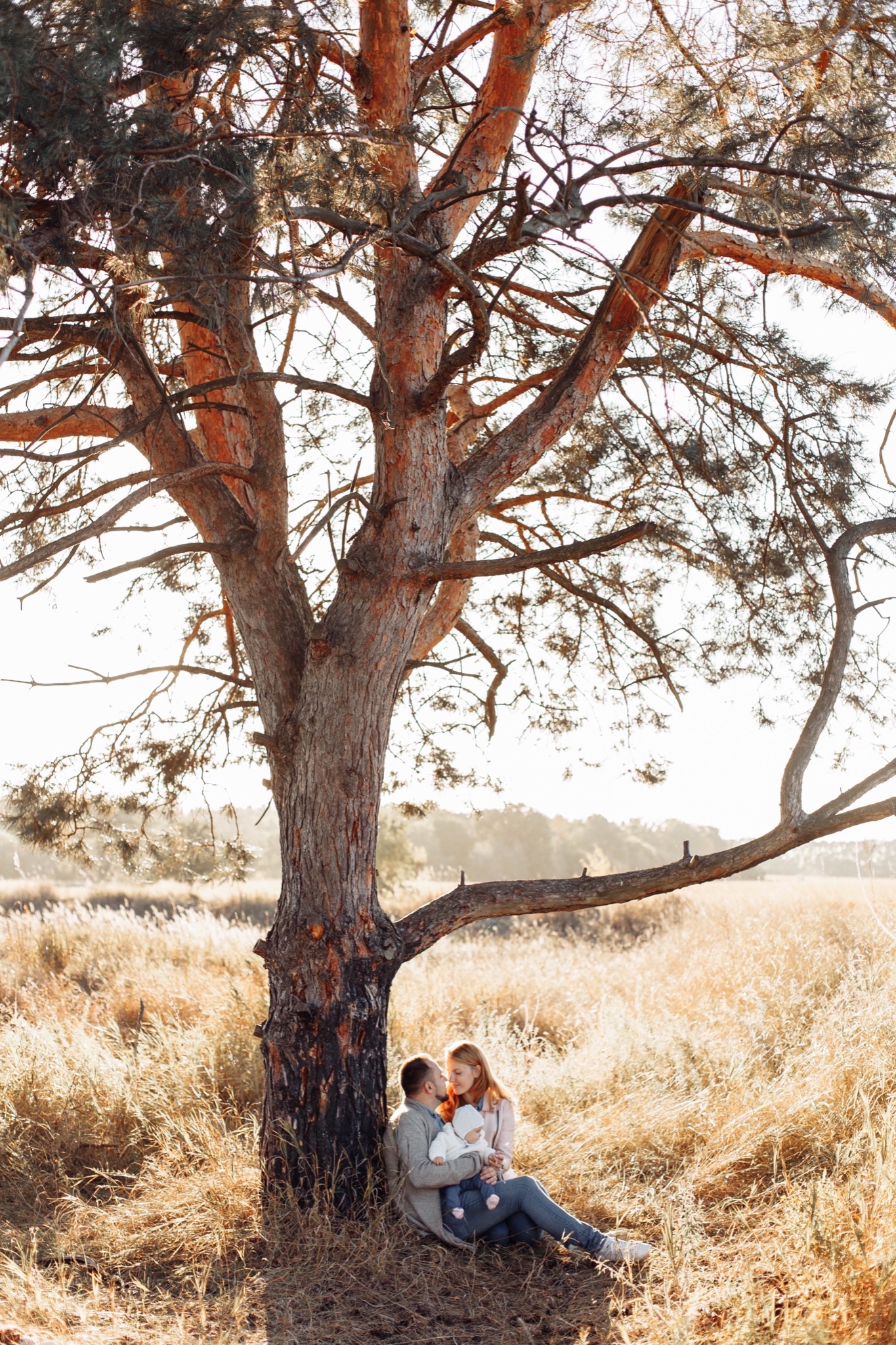 Rodzina z małym dzieckiem siedzi pod rozłożystym drzewem na tle wyschniętej trawy w słoneczny dzień.