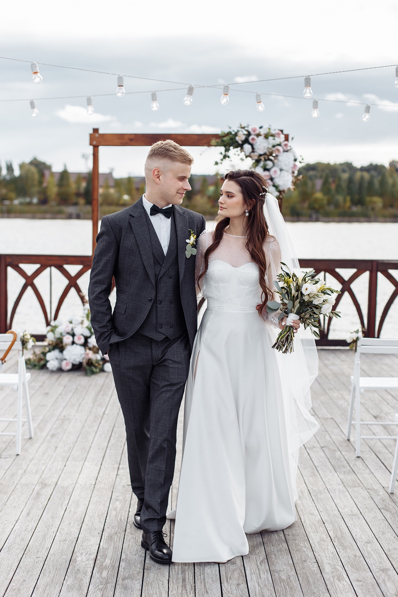 Uśmiechnięta para młoda w eleganckich strojach spaceruje po drewnianym pomoście, dekoracje kwiatowe w tle, oświetlenie żarówkami, widok na jezioro.