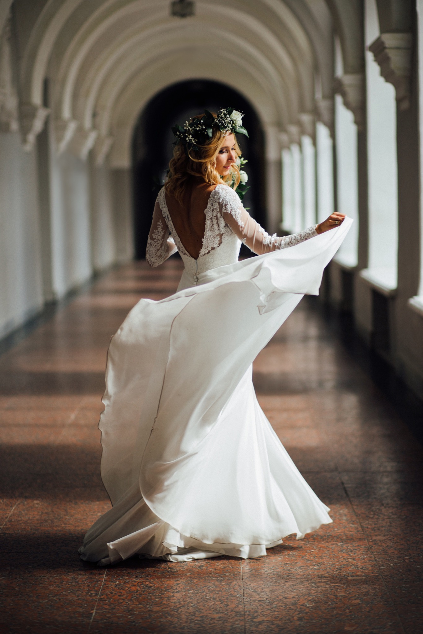Panna młoda w koronie z kwiatów, w białej sukni z koronkowymi rękawami, odwraca się w długim korytarzu z łukowatymi przejściami, unosząc brzeg sukni.